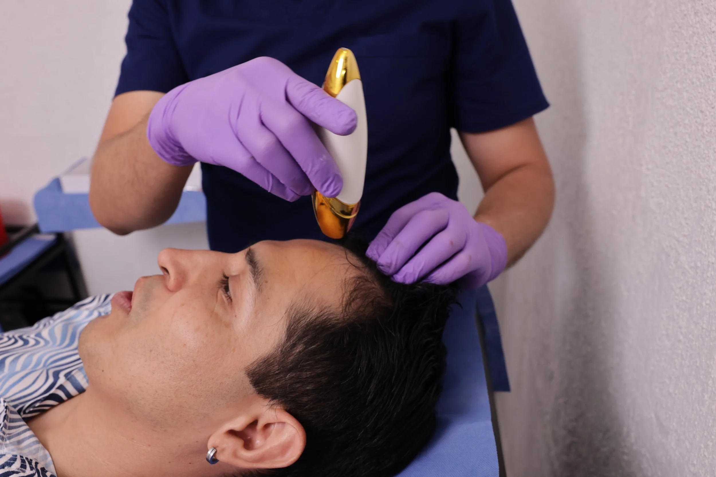 A person with short, dark hair and earrings receives cosmetic treatment from a professional wearing purple gloves, who is holding a device close to the person's face.