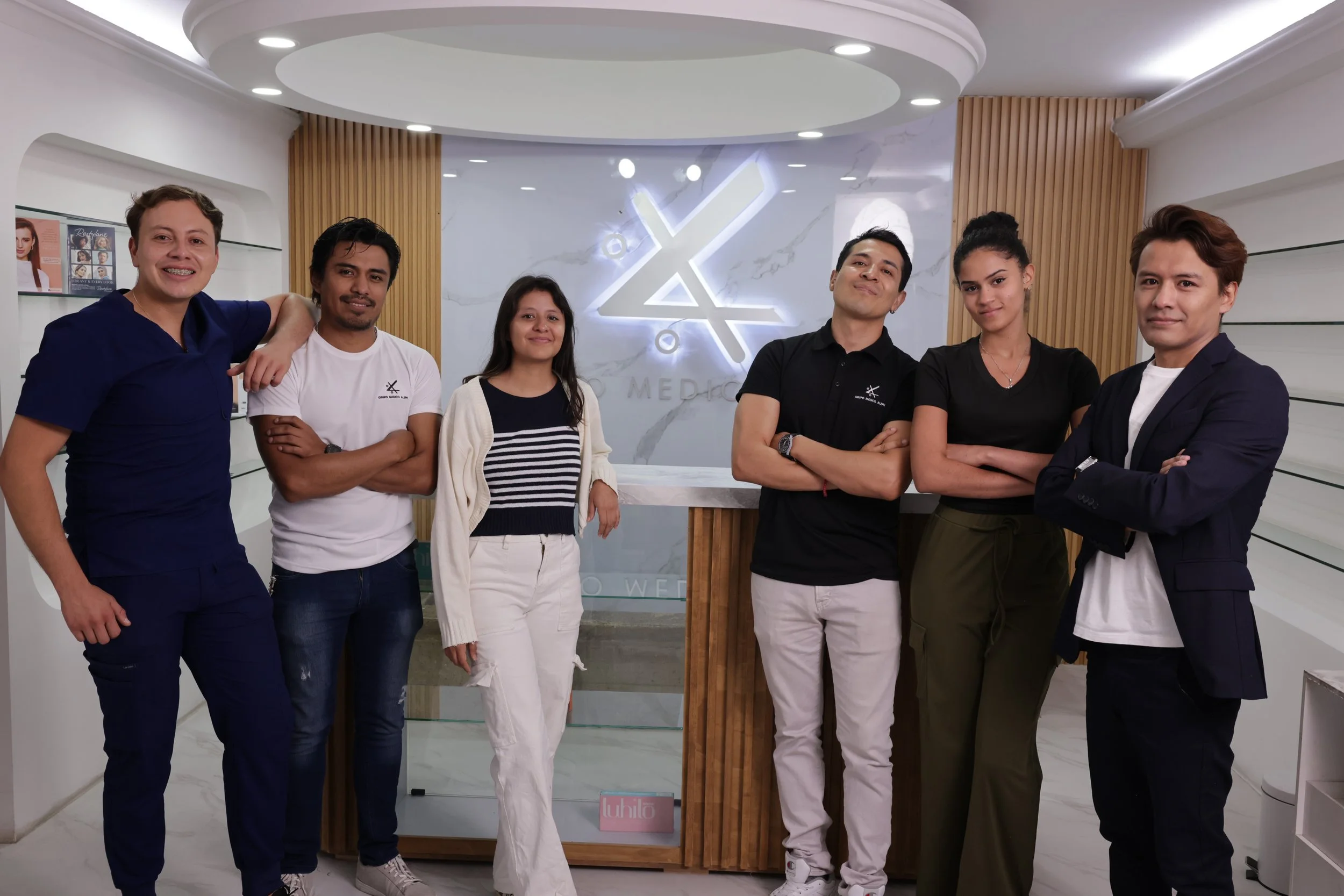 Group of six diverse people standing together in a modern medical office or clinic with a logo and wood accents in the background.