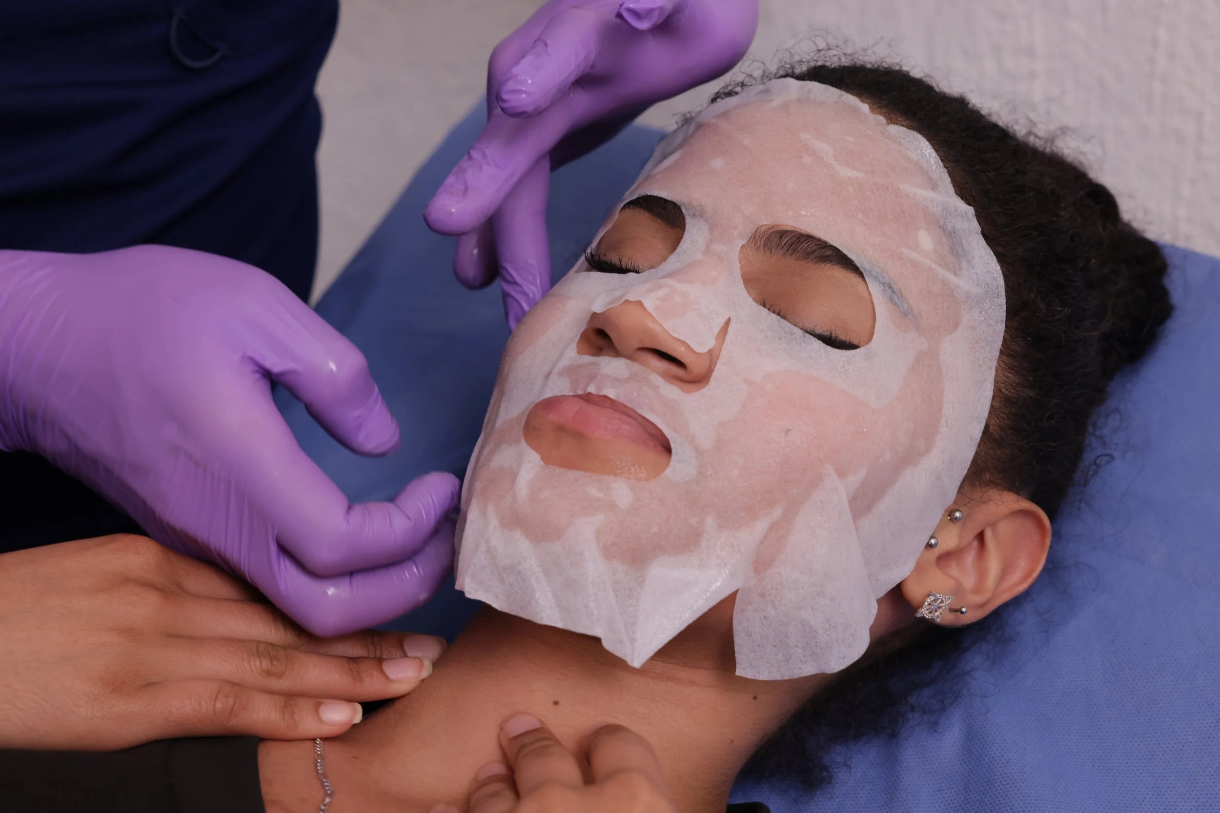 A woman receives a facial treatment while lying down with her eyes closed. A person wearing purple gloves applies a facial mask onto her face, which is covered with a sheet mask. The woman has multiple earrings in her ear and a bracelet on her wrist.
