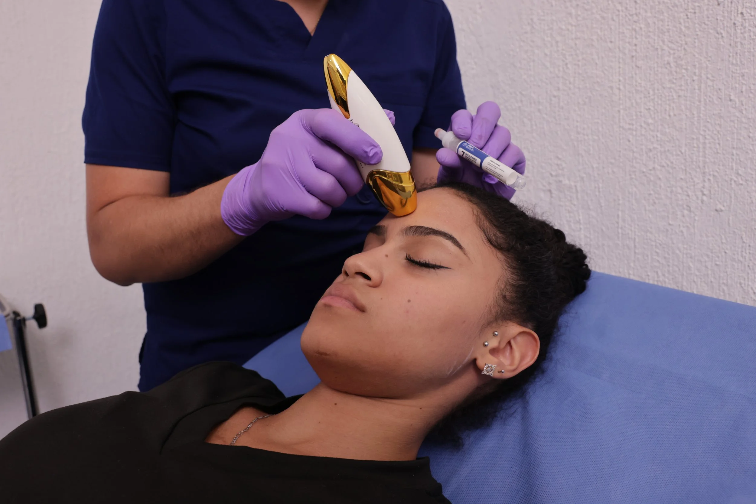 A woman with dark hair and earrings is lying down with her eyes closed while a healthcare professional, wearing purple gloves and a dark blue shirt, administers a cosmetic treatment on her forehead using a specialized device and syringe in a clinical setting with a white textured wall.
