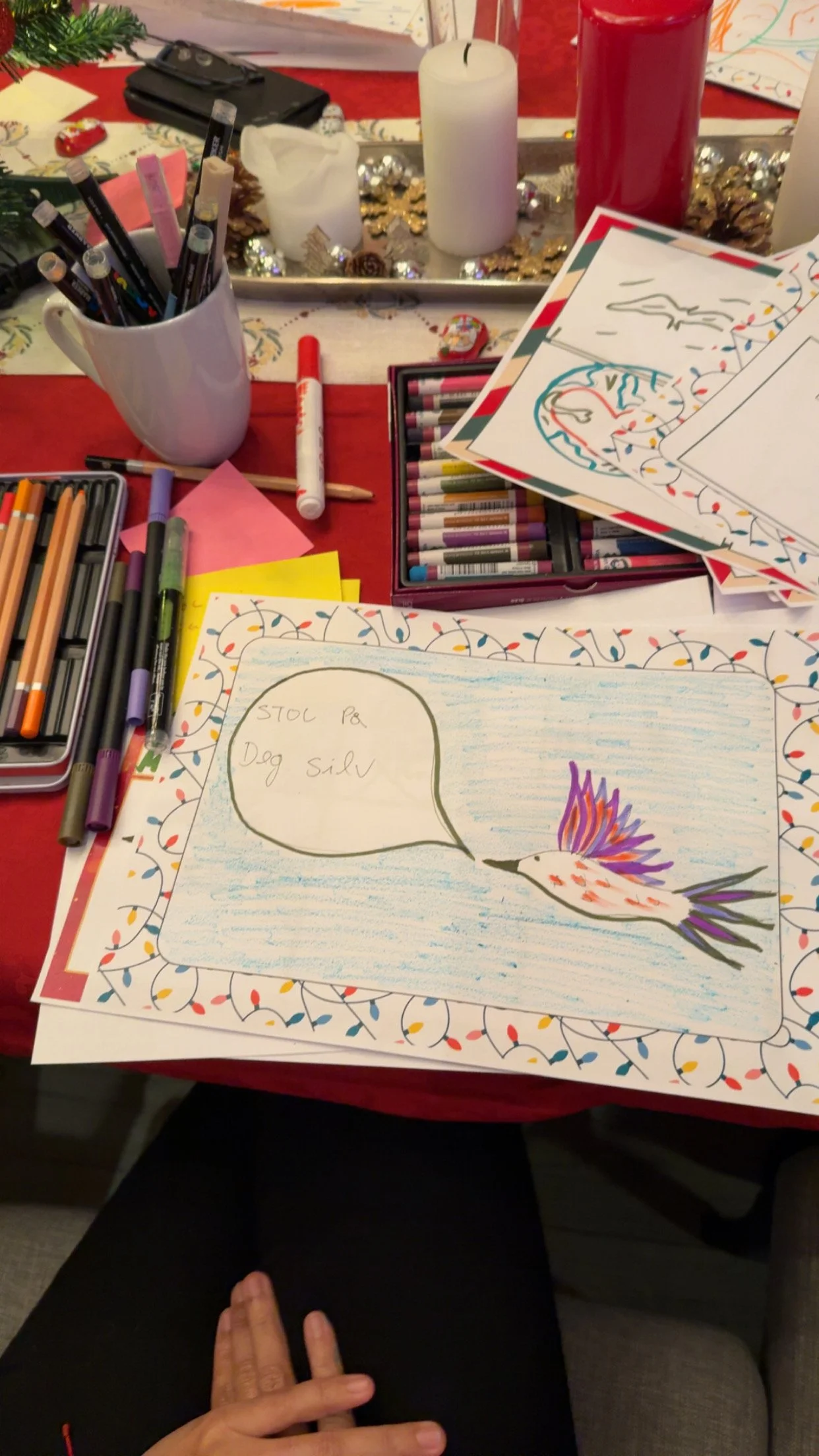 Colorful drawing of a bird with a speech bubble saying 'STOL P8. Dog Silvr' on a decorated table surrounded by markers, colored pencils, and holiday candles.