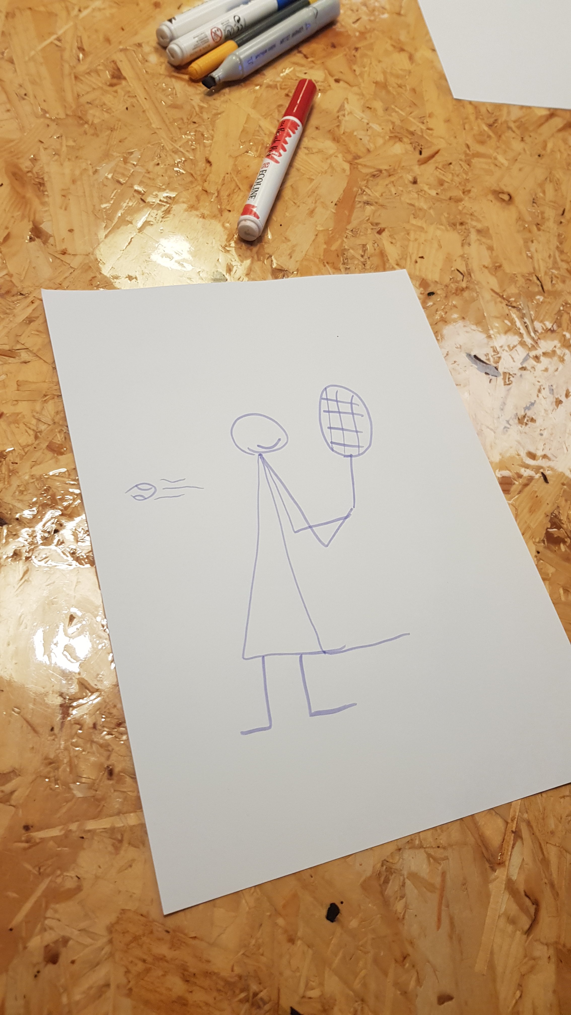 A hand-drawn stick figure girl on a white sheet of paper, holding a tennis racket, placed on a wooden surface, with several markers nearby.