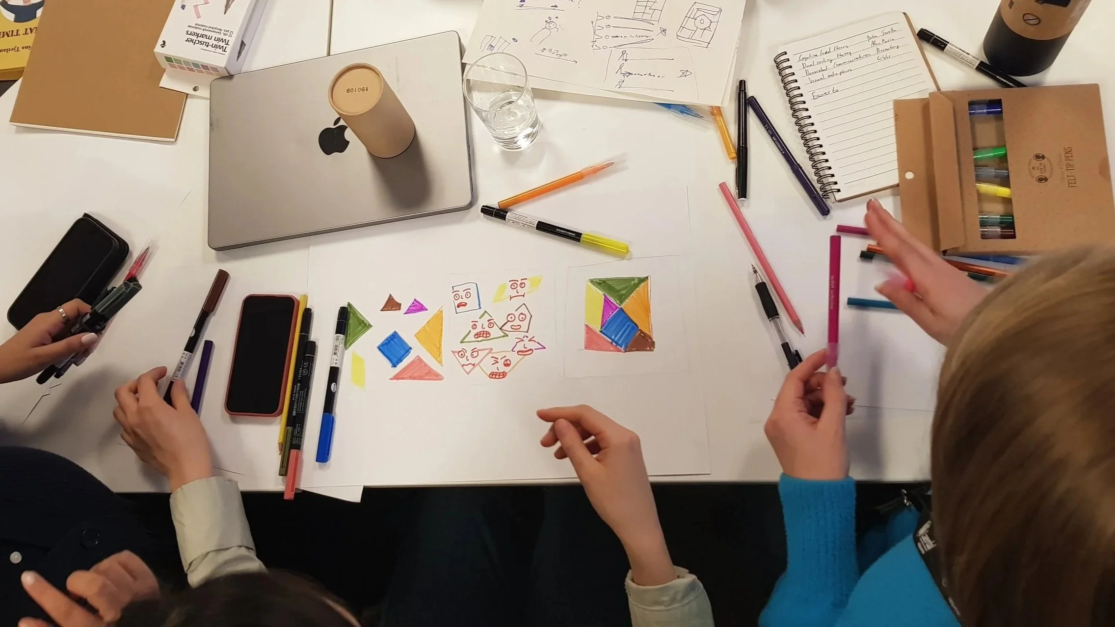 Overhead view of a group of people working on a project at a table filled with drawing papers, colored markers, pens, a closed laptop, a glass of water, a smartphone, and various sketches and colorful drawings.