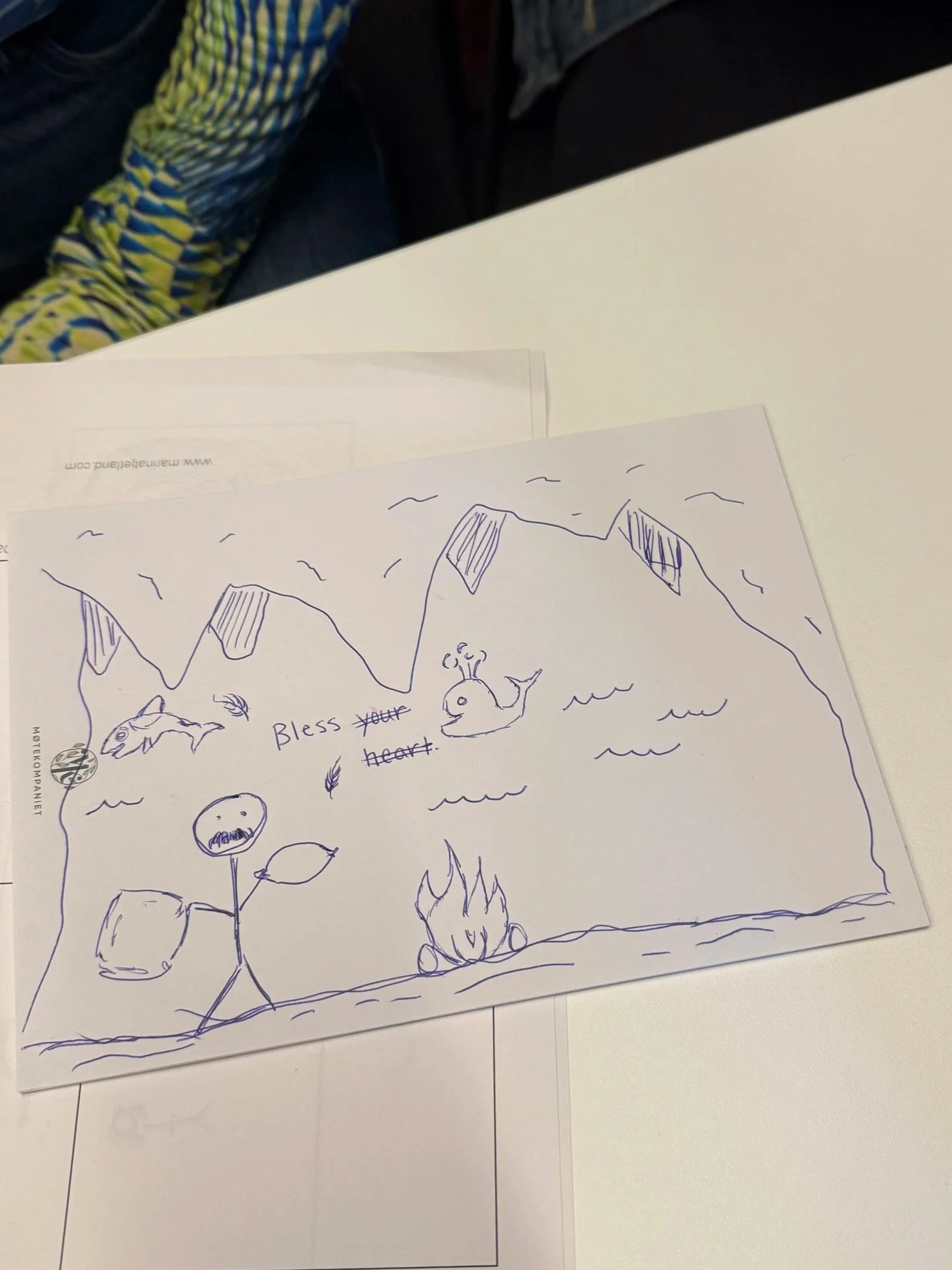A hand-drawn illustration on paper includes a whale, a whale tail, a fish, a flower, and the words 'Bless your heart' written on the paper. The drawing is in simple blue ink and is placed on a white table.