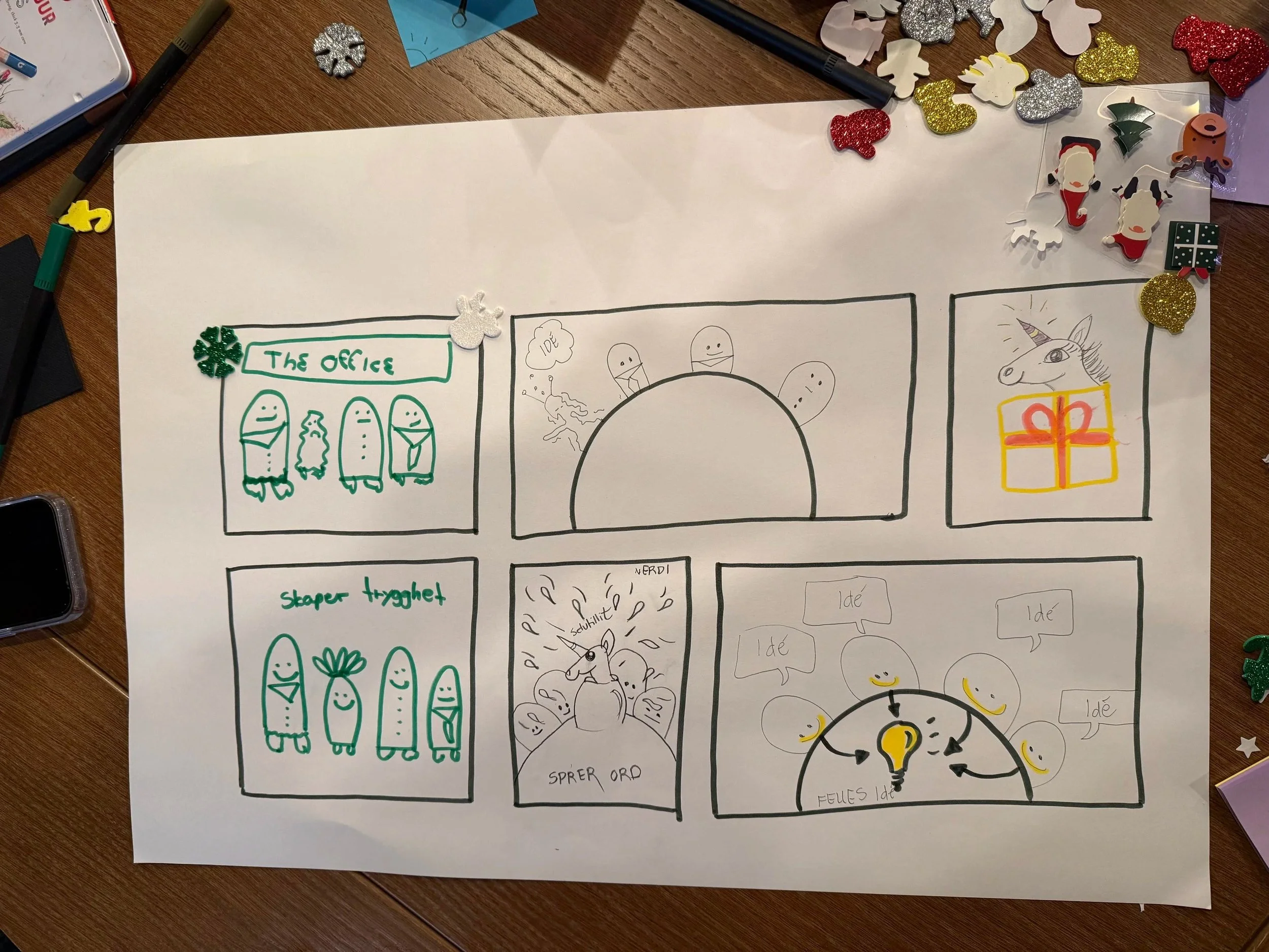 A handwritten comic strip on a large sheet of paper surrounded by various decorative items including glittery snowflakes, Christmas-related embellishments, and marker pens. The comic appears to involve illustrations of characters and text, with some 