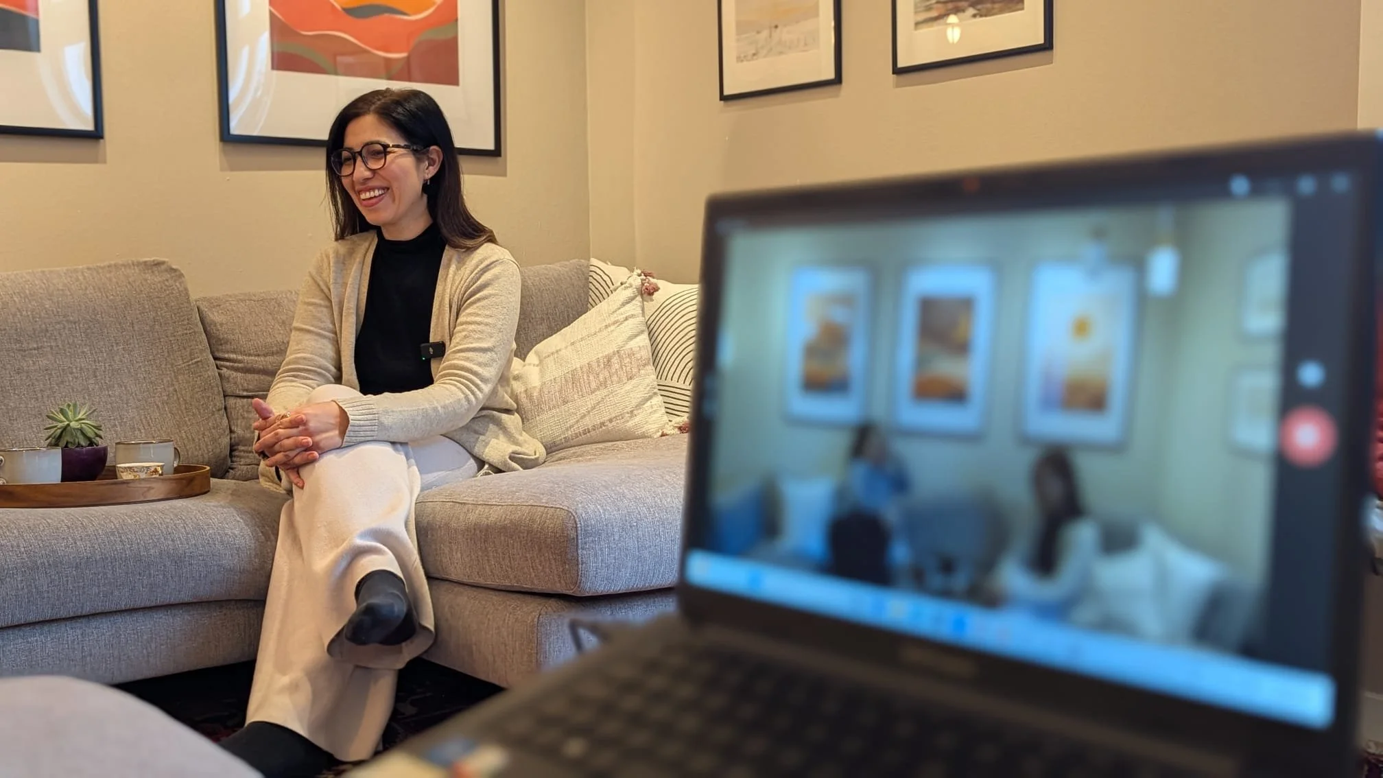 A woman with dark hair, glasses, and a beige cardigan sitting on a gray sofa, smiling for a photo during an indoor interview or recording session.