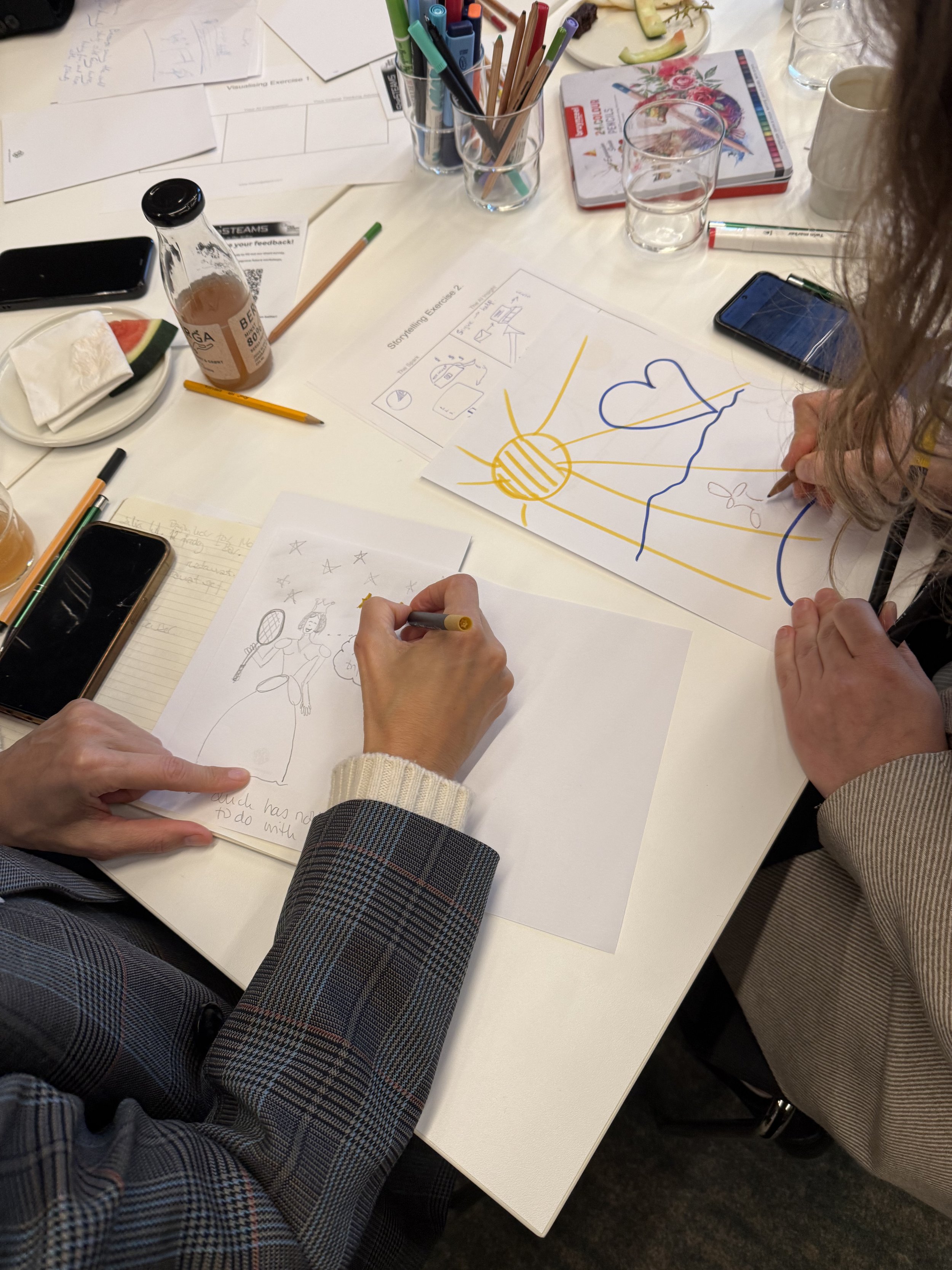 People sitting around a table with drawing papers, markers, notebooks, drinks, and snacks, engaged in a creative or brainstorming activity.