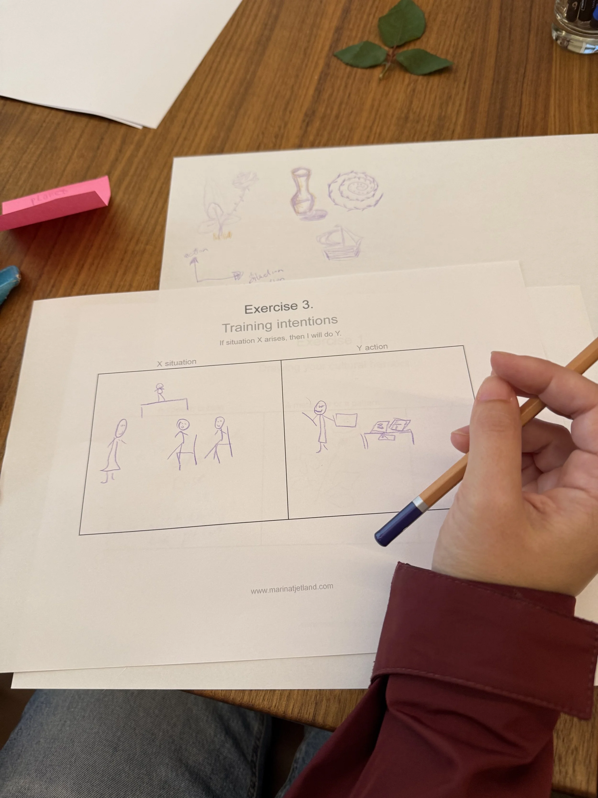 Person holding a pen working on a worksheet titled "Exercise 3. Training intentions" with sketches and notes on a table.