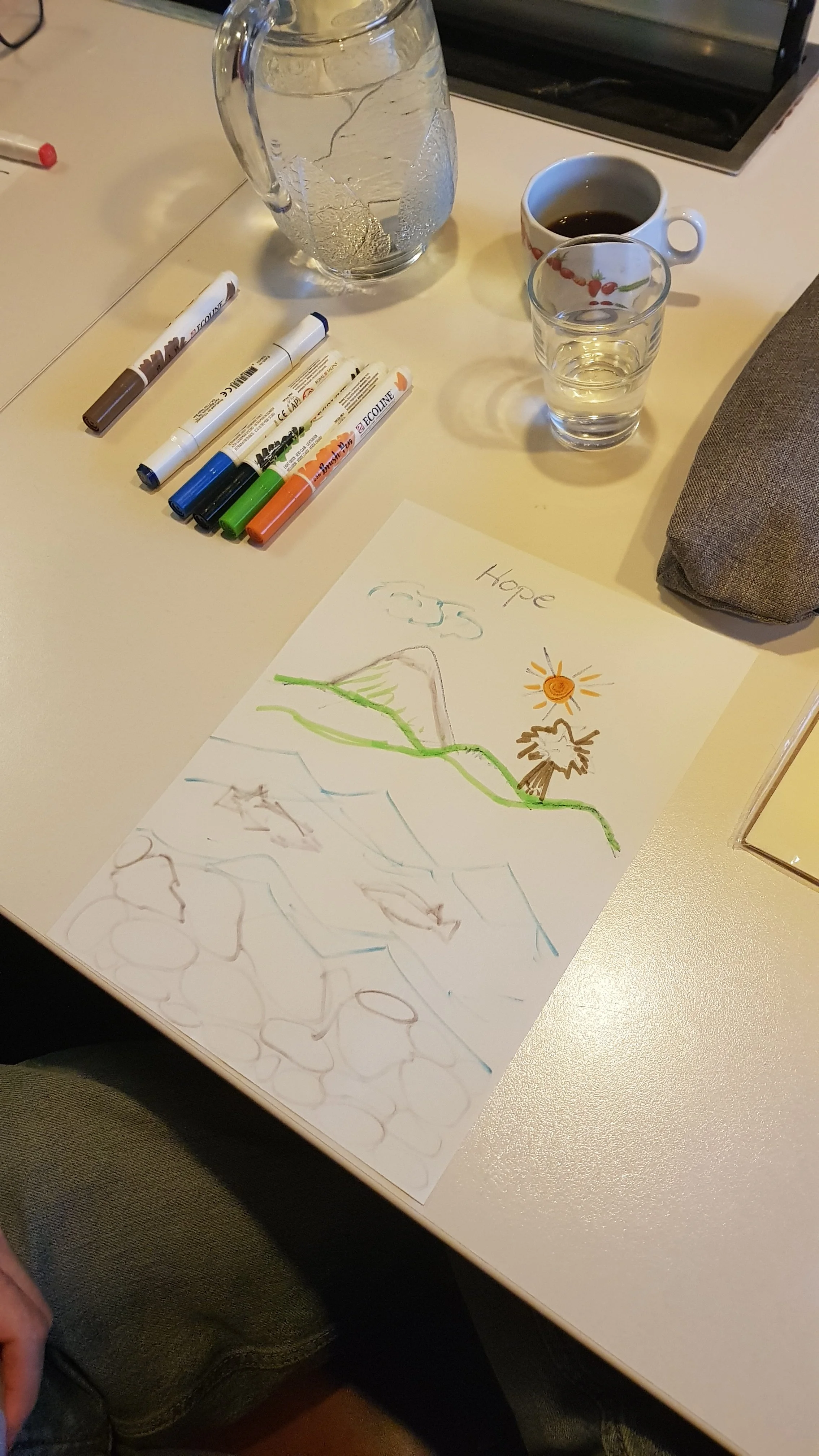 A piece of paper with a child's colorful drawing of a mountain, a tree, clouds, and a shining sun, with the word "Hope" written at the top. Around the paper are markers, a glass of water, a cup of coffee, and a water pitcher on a beige table.