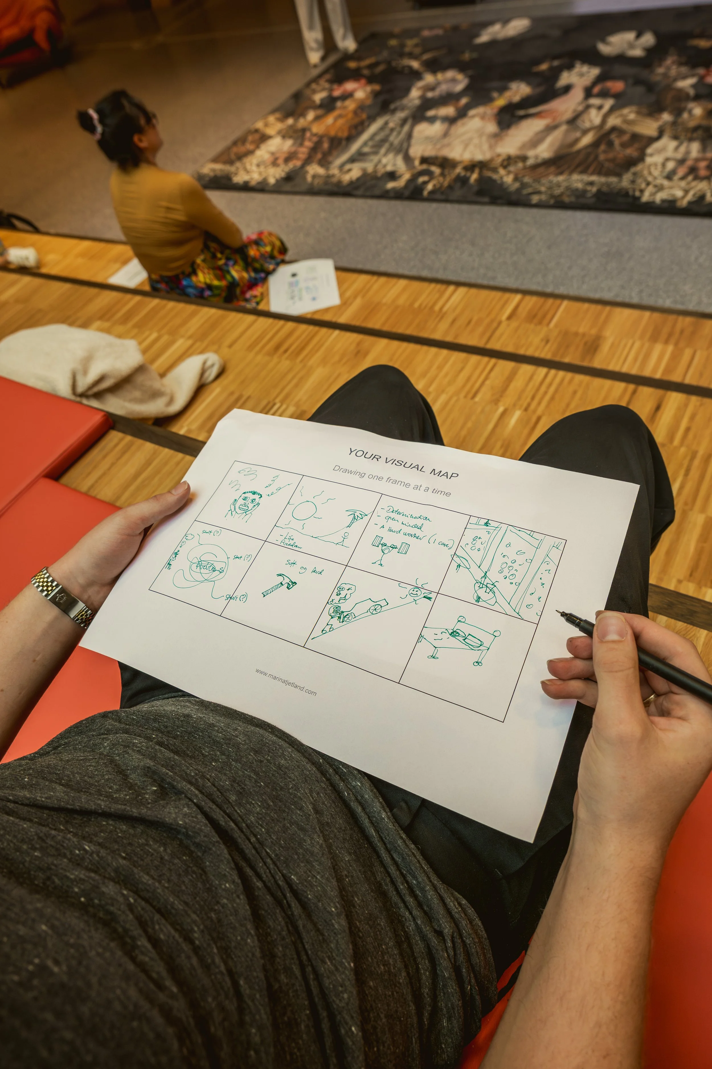 A person holding a drawing sheet with sketches, sitting in a room, with a child sitting on the floor in front of an artwork. The person has a pen in hand, and the sketch sheet has a grid of doodles and ideas.