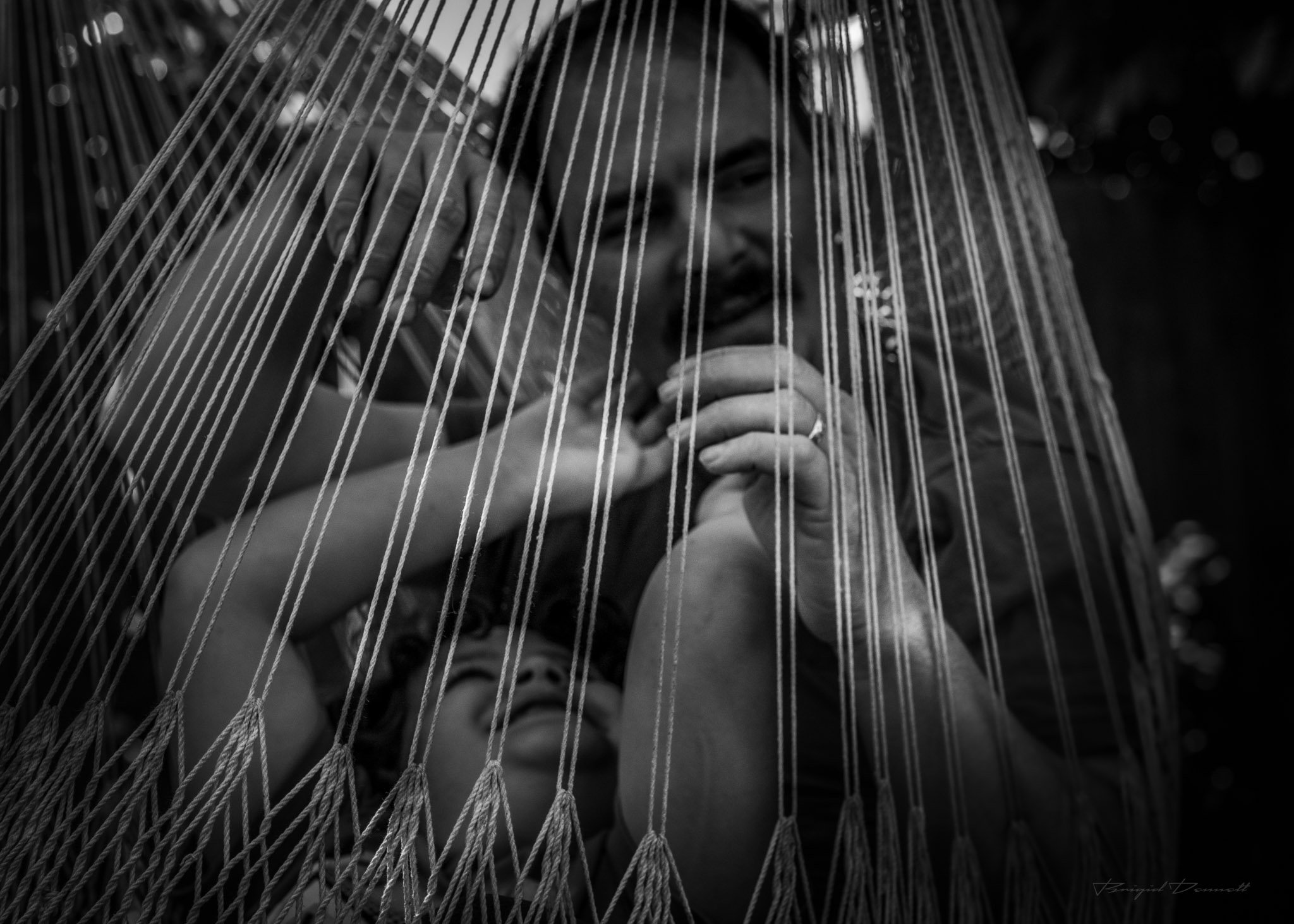 A person sitting on a hammock, viewed through the hammock's strings, drinking from a cup.