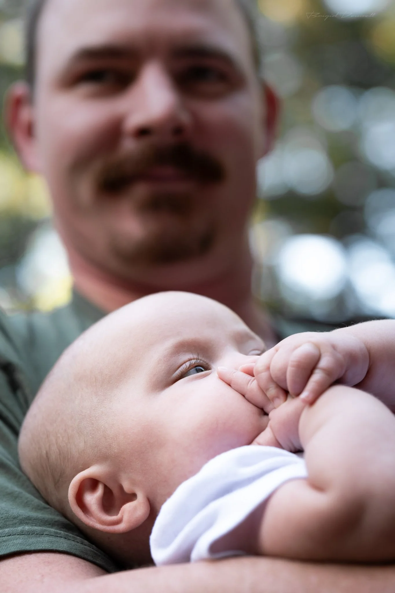 A man with a mustache holding a baby close, the baby's hand touching his face, outdoors with blurred natural background.