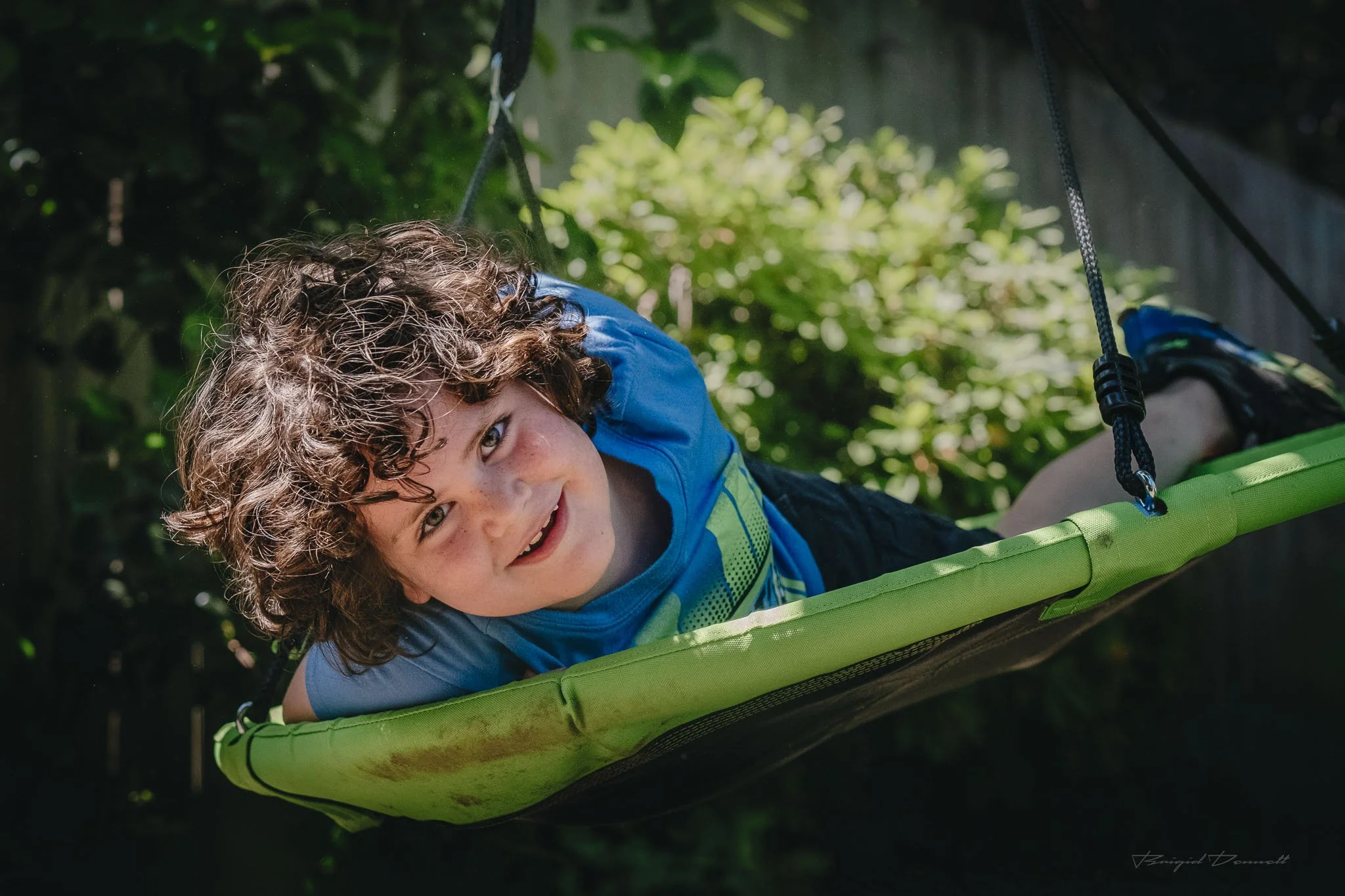Young boy with curly hair smiling while lying on a green outdoor swing in a garden.