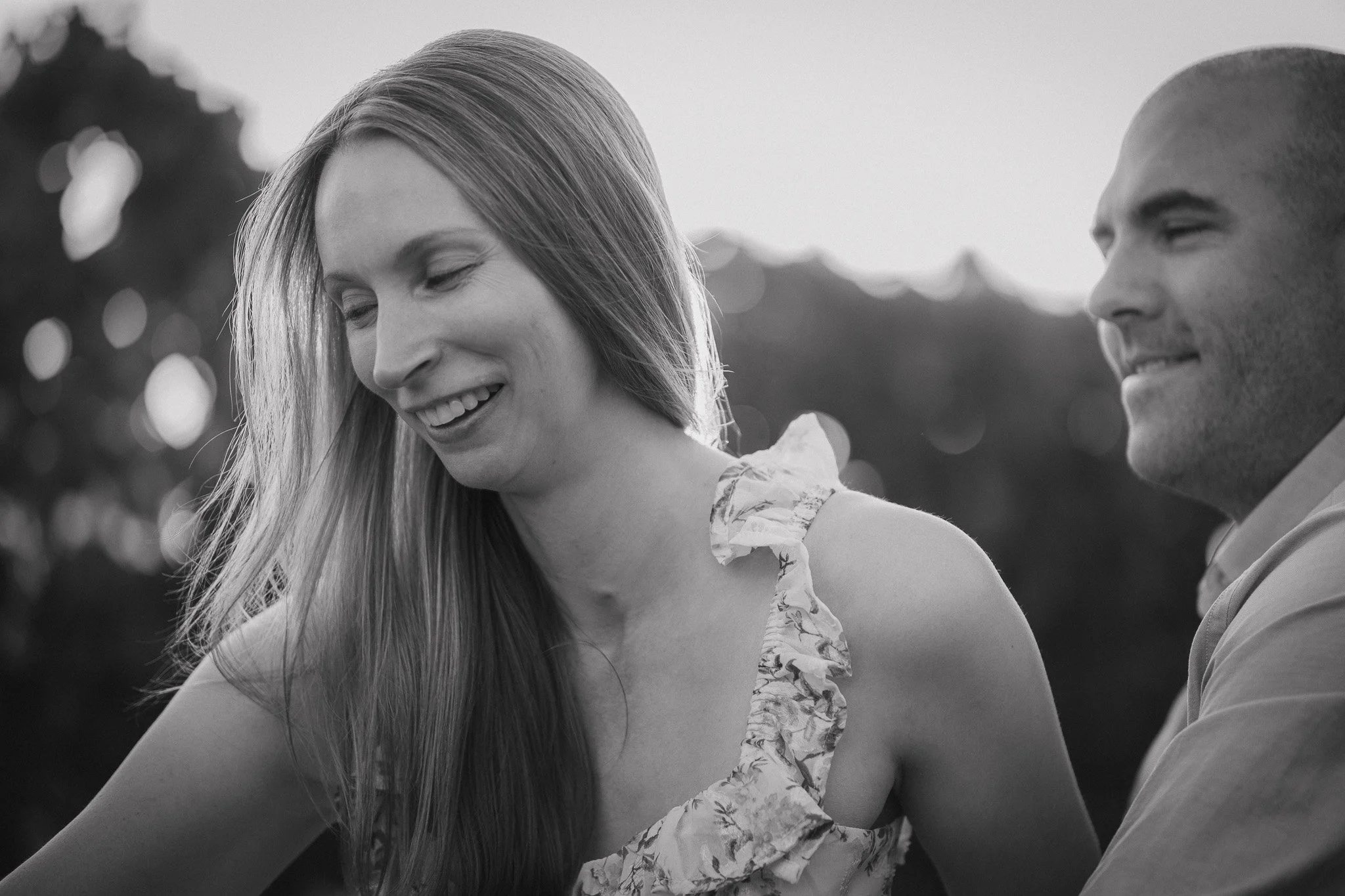 Black and white photo of a man and woman smiling and looking at each other, outdoors during sunset or evening.
