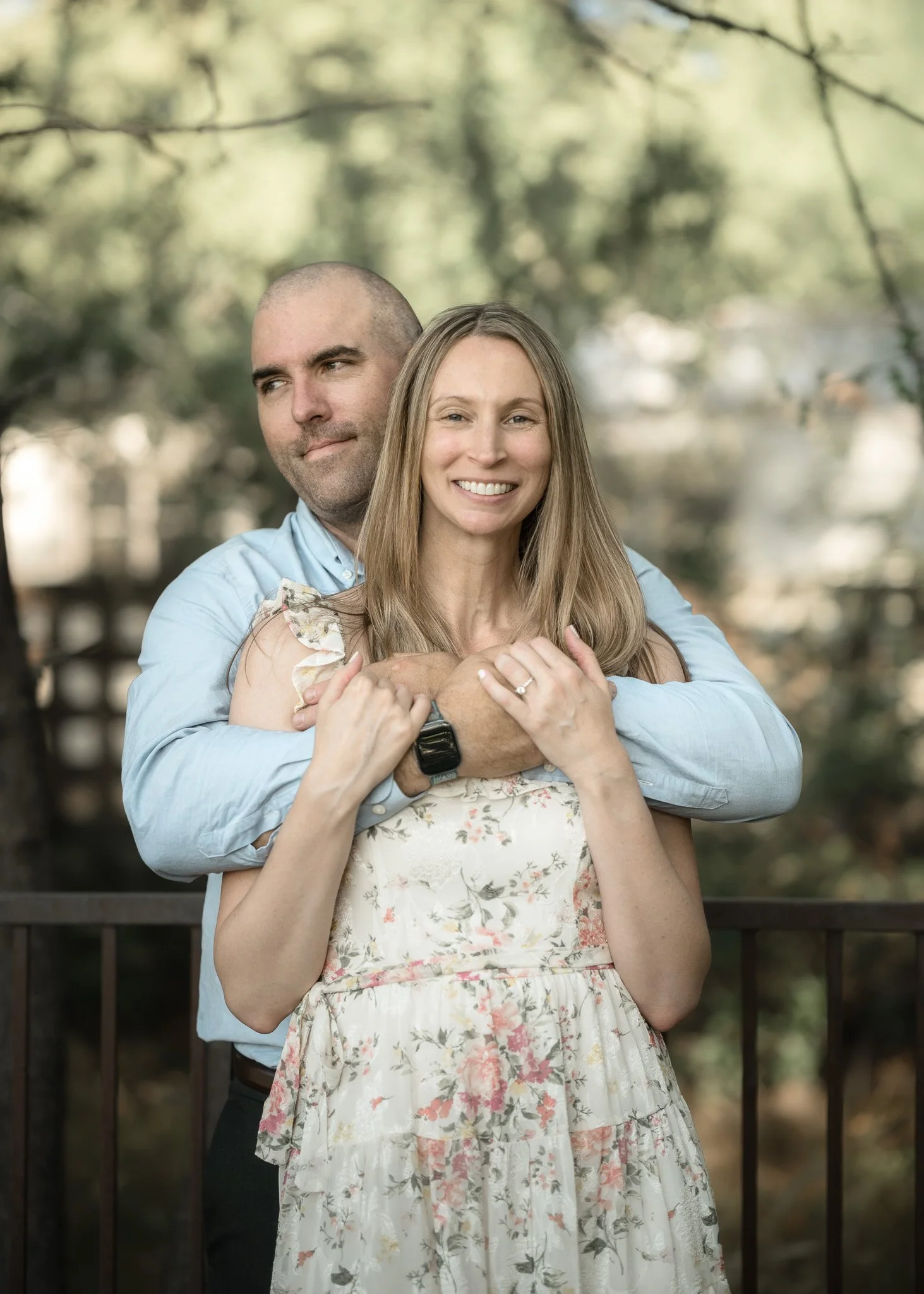 A smiling woman with long blonde hair standing in front of a man with a shaved head, both embracing each other outdoors in front of a fence and trees.