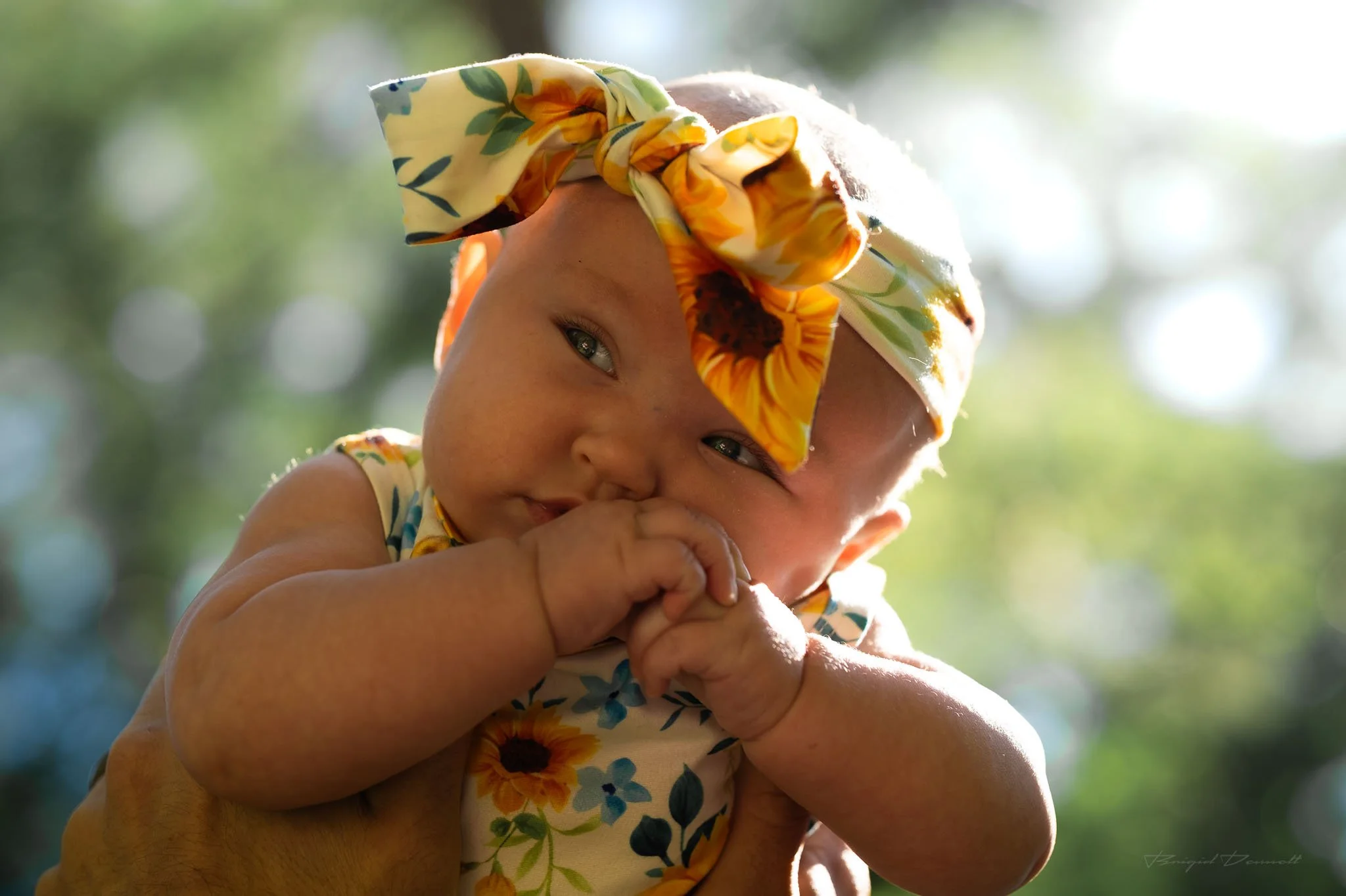 A baby with a floral headscarf being held up outdoors, with trees and sunlight in the background.