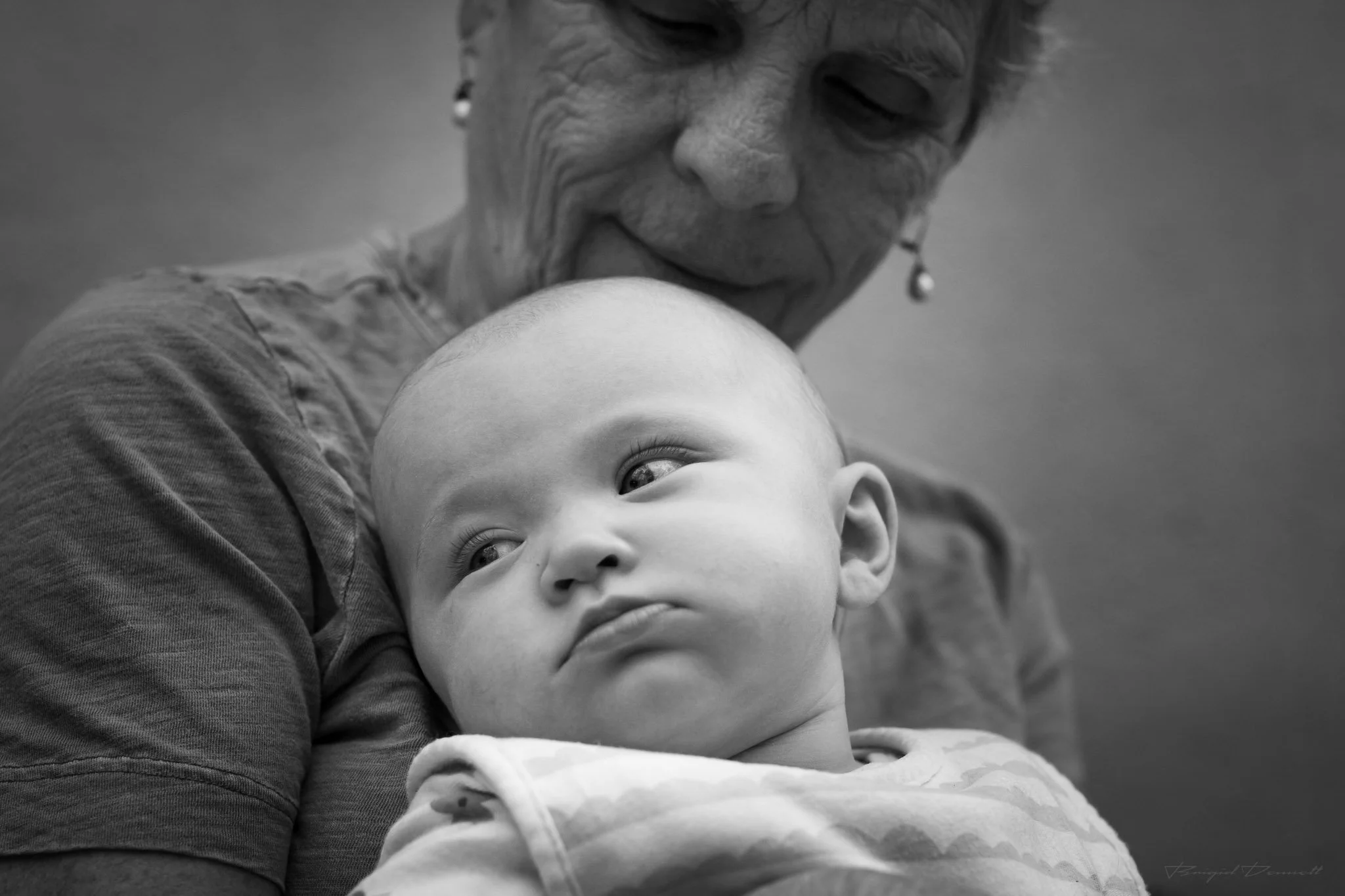 A black and white close-up photograph of an elderly woman holding a baby. The woman has her eyes closed, wearing earrings, and seems to be in a moment of affection or contemplation. The baby, resting in the woman's arms, has big eyes looking slightly