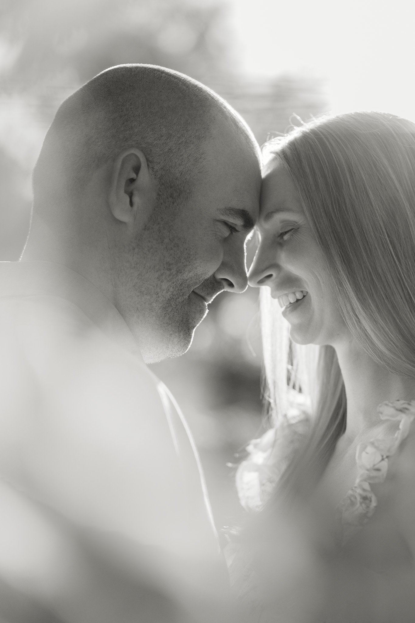 A black and white photo of a couple with their foreheads touching, smiling, with closed eyes, outdoors with sunlight shining behind them.