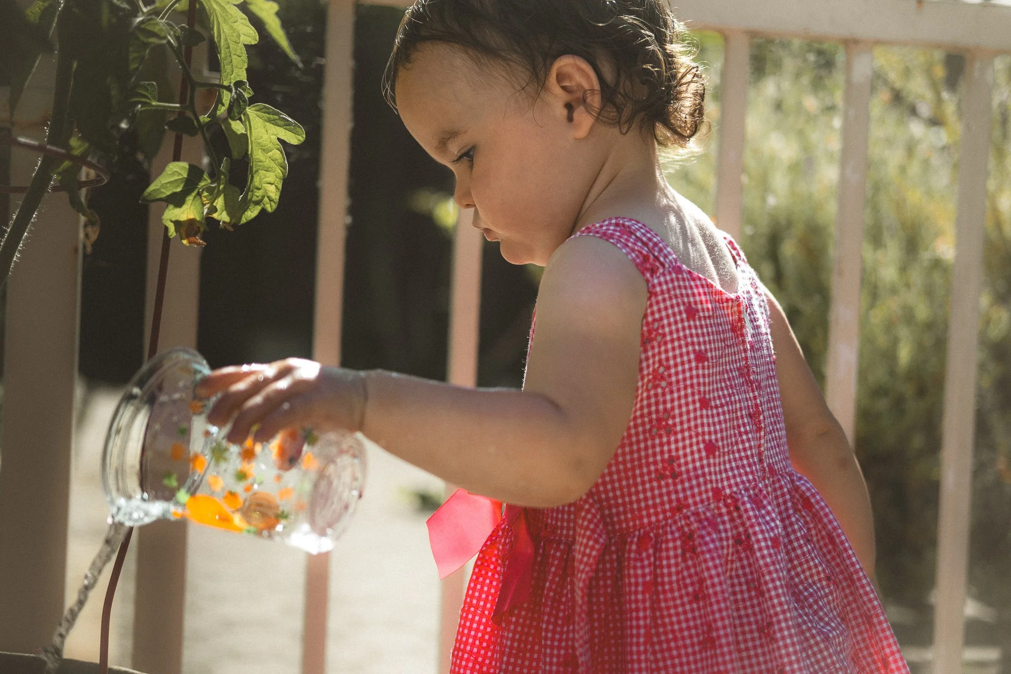 A young girl in a red checkered dress is outdoors, watering flowers in a glass jar on a porch or balcony, with sunlight highlighting her.