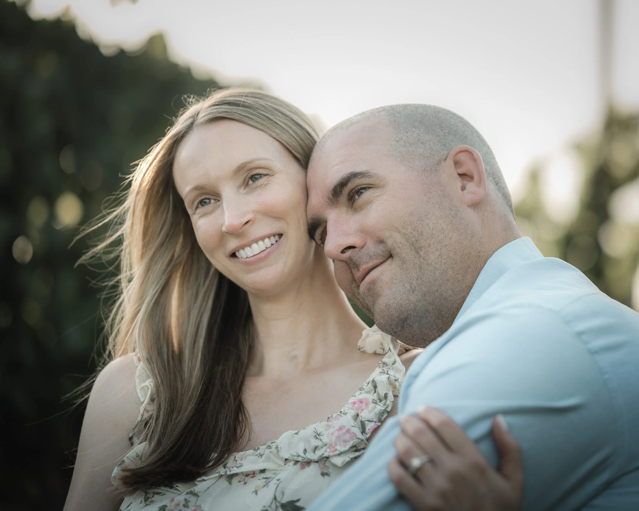 A smiling woman with long blonde hair and a man with a shaved head, dressed in light-colored clothing, leaning close together outdoors with blurred greenery in the background.