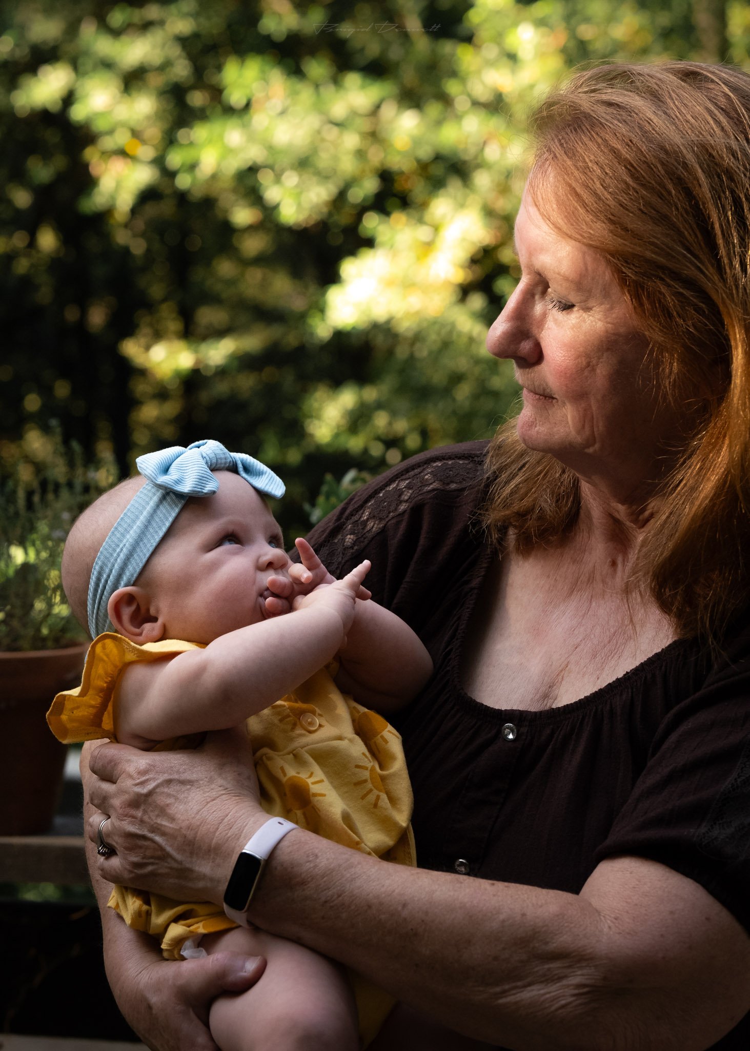 An elderly woman with long red hair holding a baby girl with a blue bow headband outdoors. The woman has her eyes closed and the baby is looking up at her.