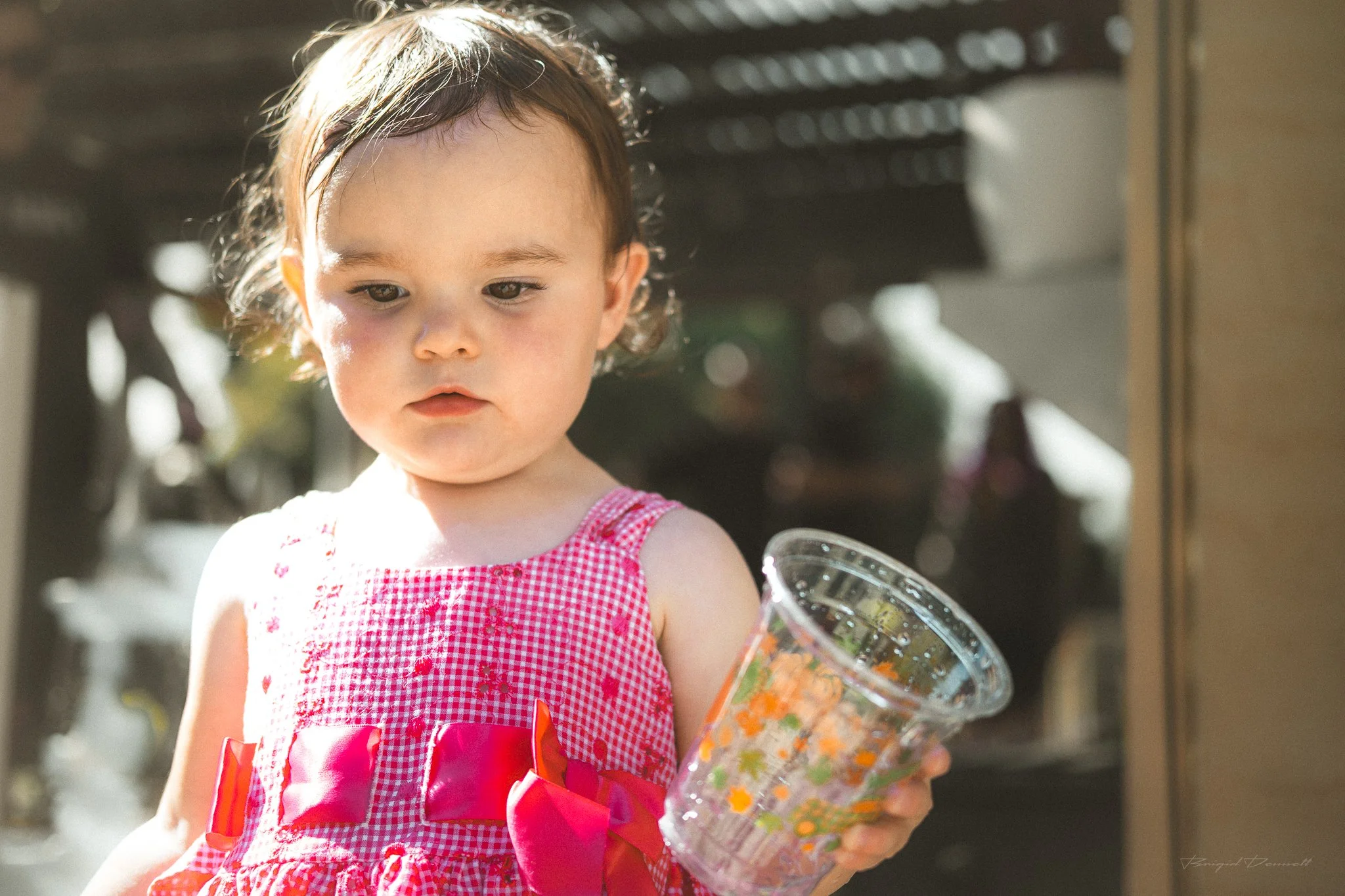 A young girl with brown hair in a ponytail, wearing a red and white checkered dress with a red bow, holding a container of colorful snacks or candy outside.