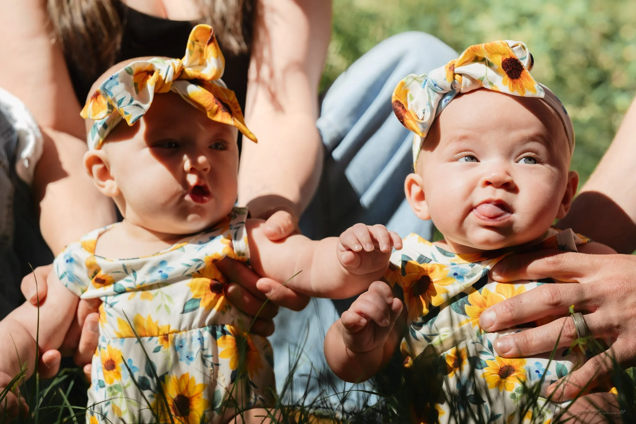 Two babies wearing sunflower-patterned headbands and dresses, being held outdoors with a blurred green background.