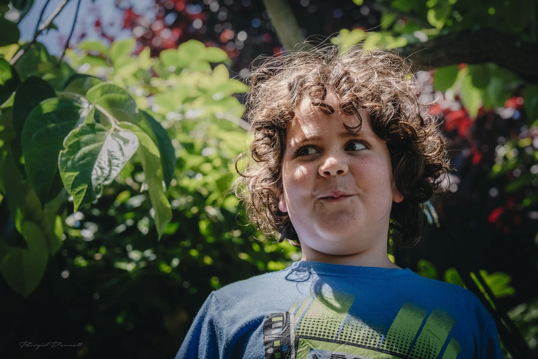 A young boy with curly hair looks to the side outdoors among green leaves.