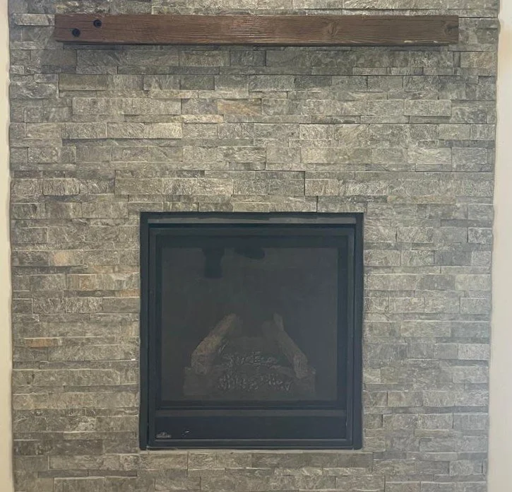 Interior of a building with a stone fireplace at the center, flanked by two open doorways, with dark wood flooring, textured white ceiling with recessed lighting, and walls covered with small mosaic tiles.