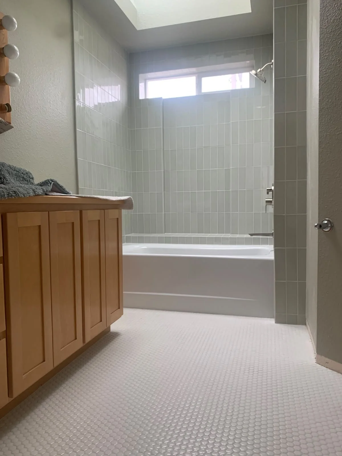 Modern bathroom with gray tiled walls and floor, a white wall-mounted sink with a wooden countertop, a round mirror, a black soap dispenser, a white towel radiator on the wall, and ceiling lights.