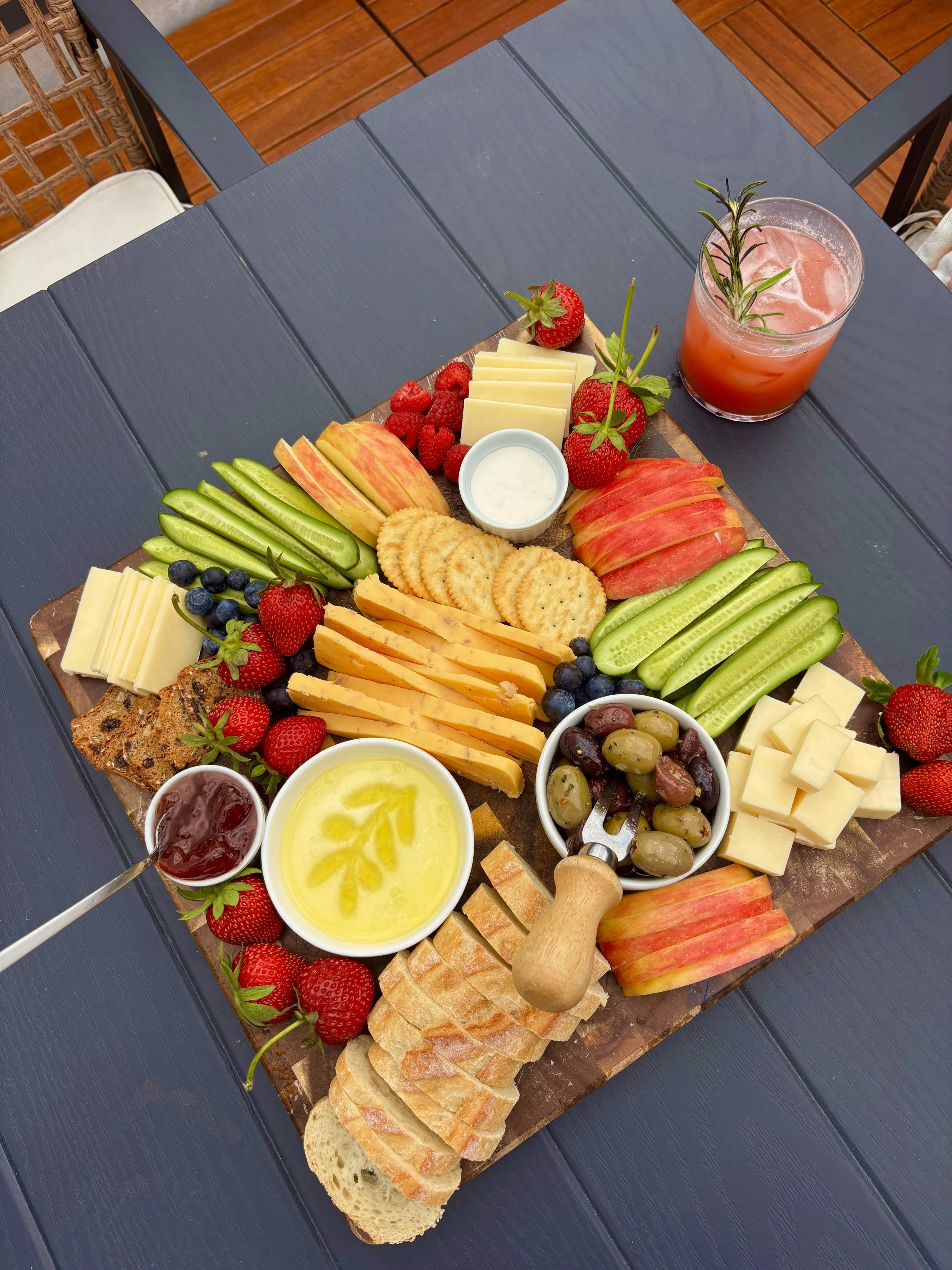 A cheese and fruit platter on a wooden board featuring sliced apples, cucumbers, strawberries, blueberries, raspberries, cheese slices, crackers, and dipping sauces, accompanied by a glass of pink beverage garnished with rosemary on a dark wooden table.