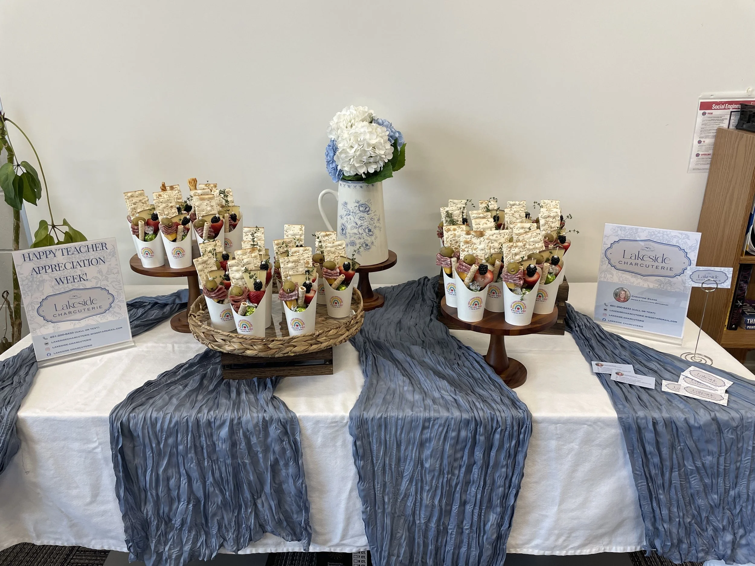 Display table with decorative cupcakes in paper cups, a vase with white and blue flowers, and informational signs for Lakeside Charcuterie and Teacher Appreciation Week.
