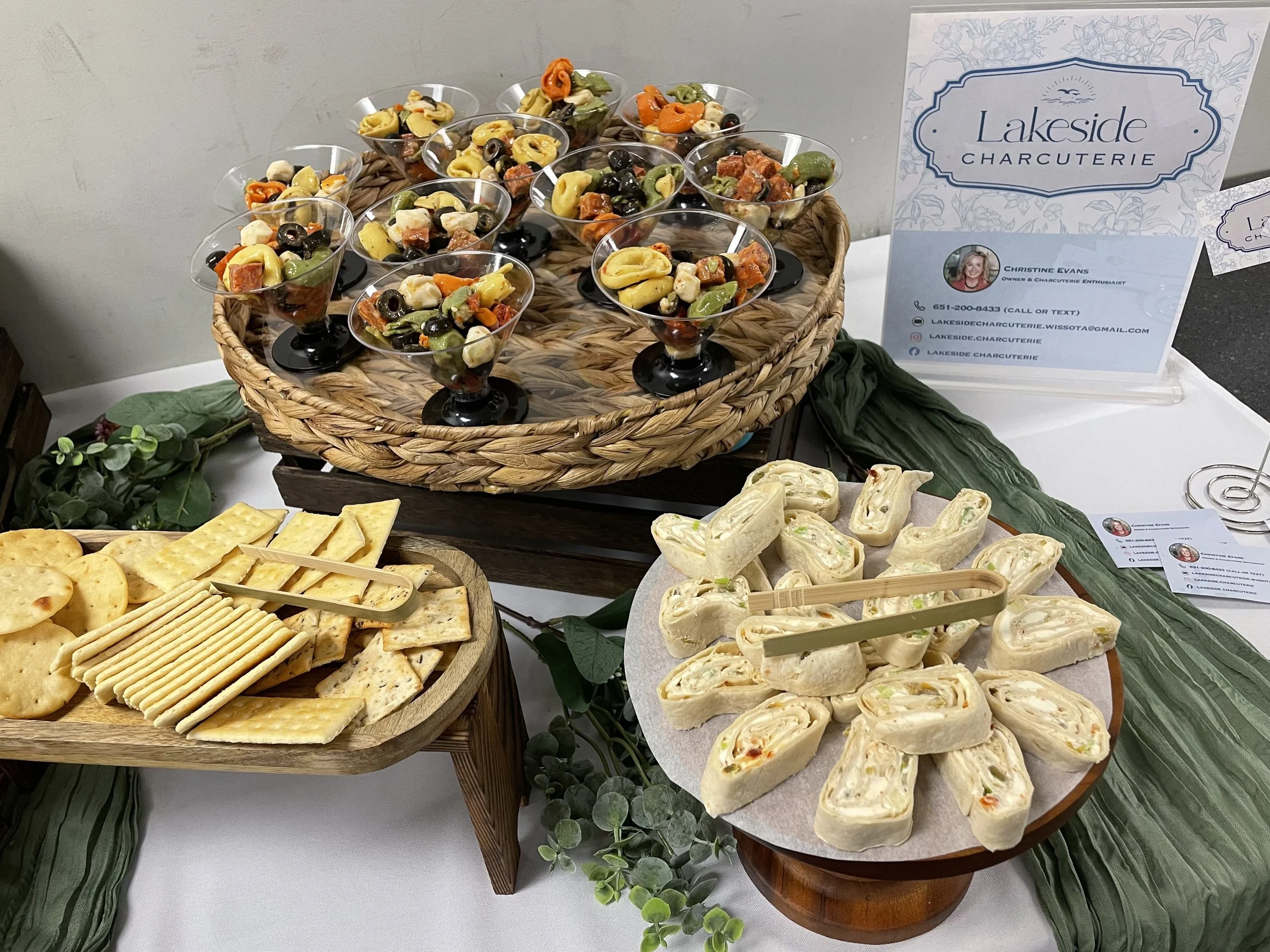 A platter of sliced wraps filled with vegetables and possibly some protein, displayed on a table alongside crackers, cheese, and other appetizers. The table has a greenery decoration, and there are informational signs for Lakeside Charcuterie.