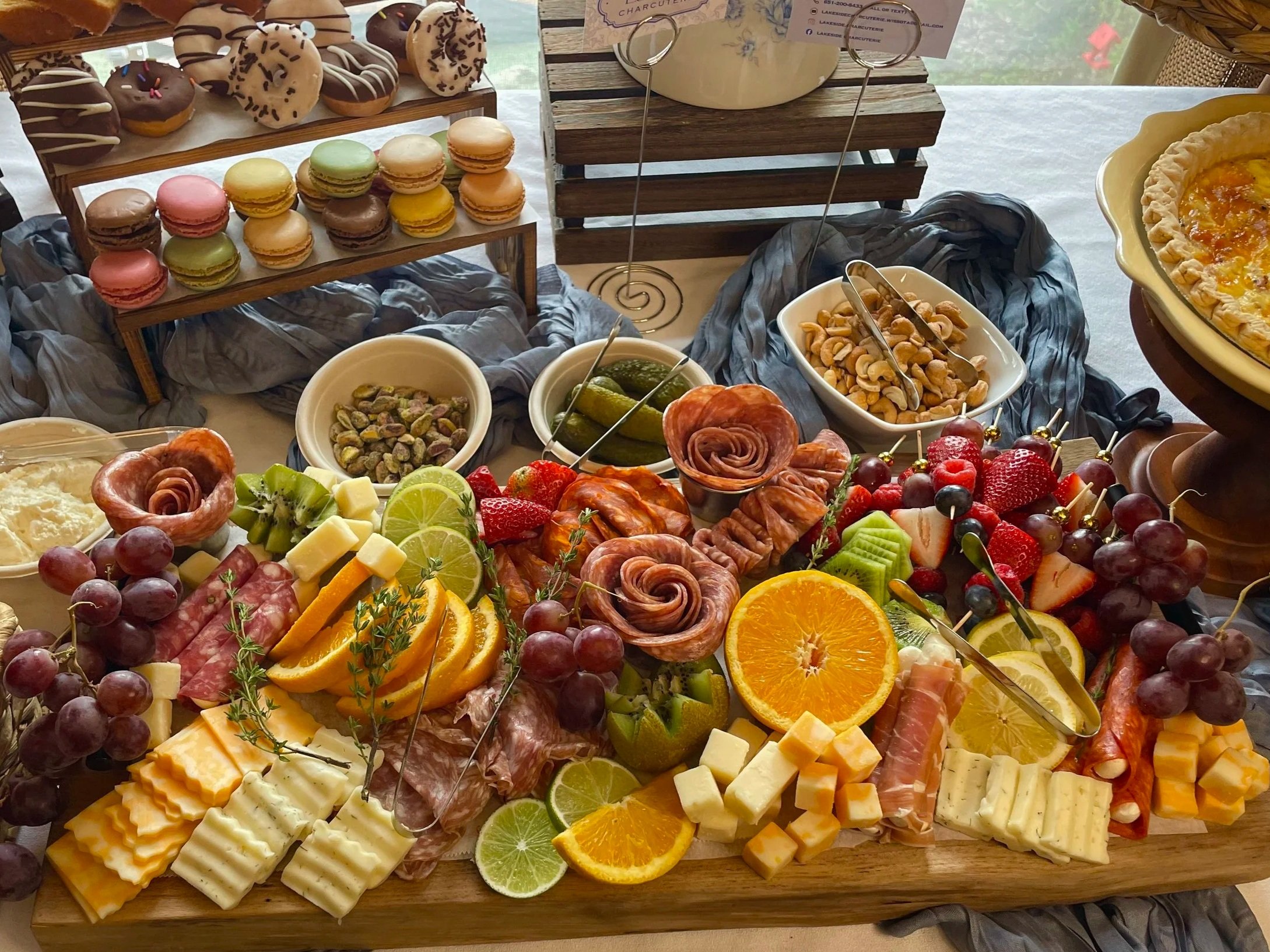 Display of various desserts and snacks including macarons, donuts, cheeses, fruits, nuts, and cured meats on a table at a buffet or party.