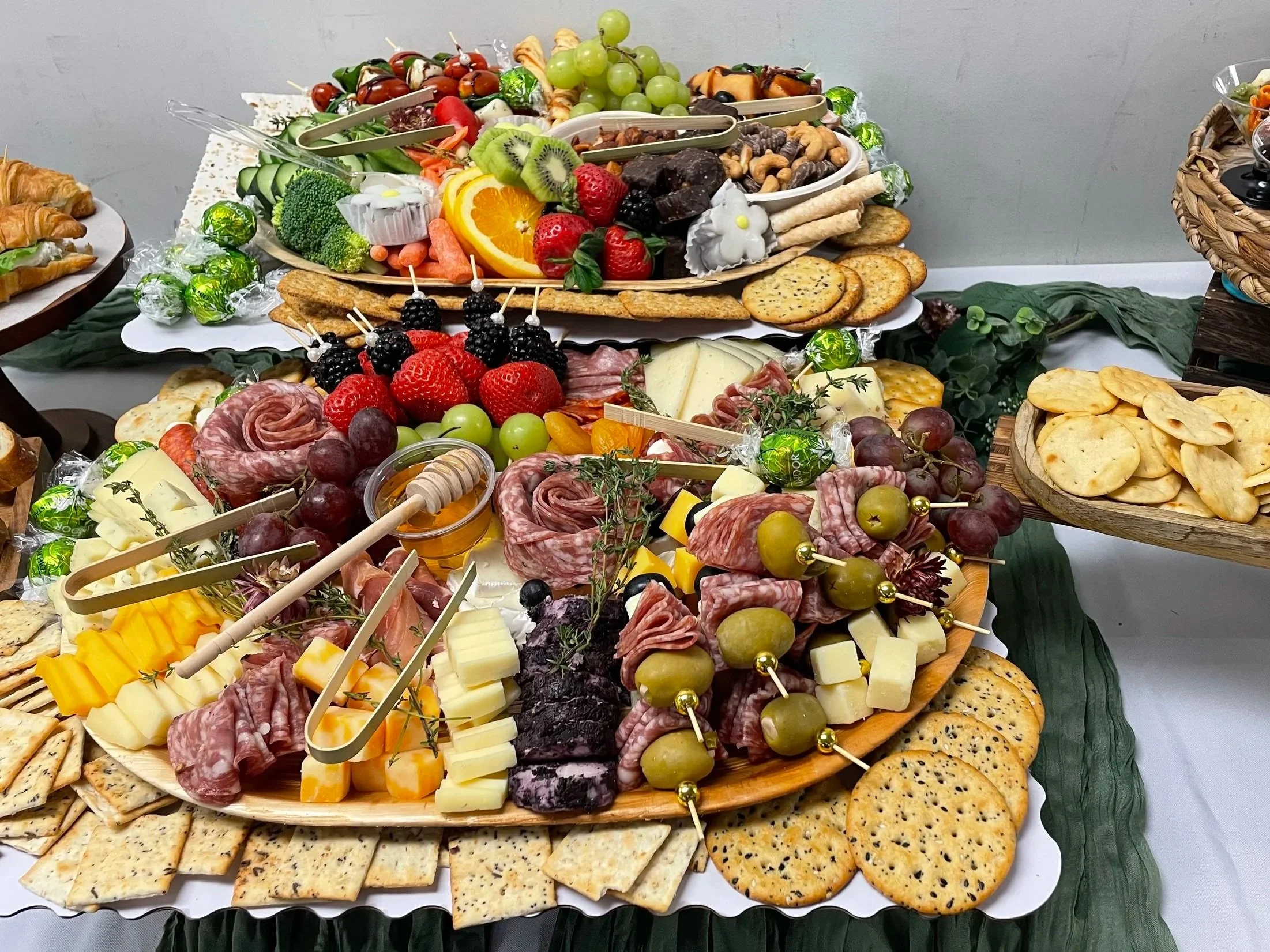 An elaborate cheese and charcuterie board with crackers, grapes, strawberries, kiwi, oranges, cherry tomatoes, broccoli, and assorted meats and cheeses, garnished with decorative flowers and greenery.