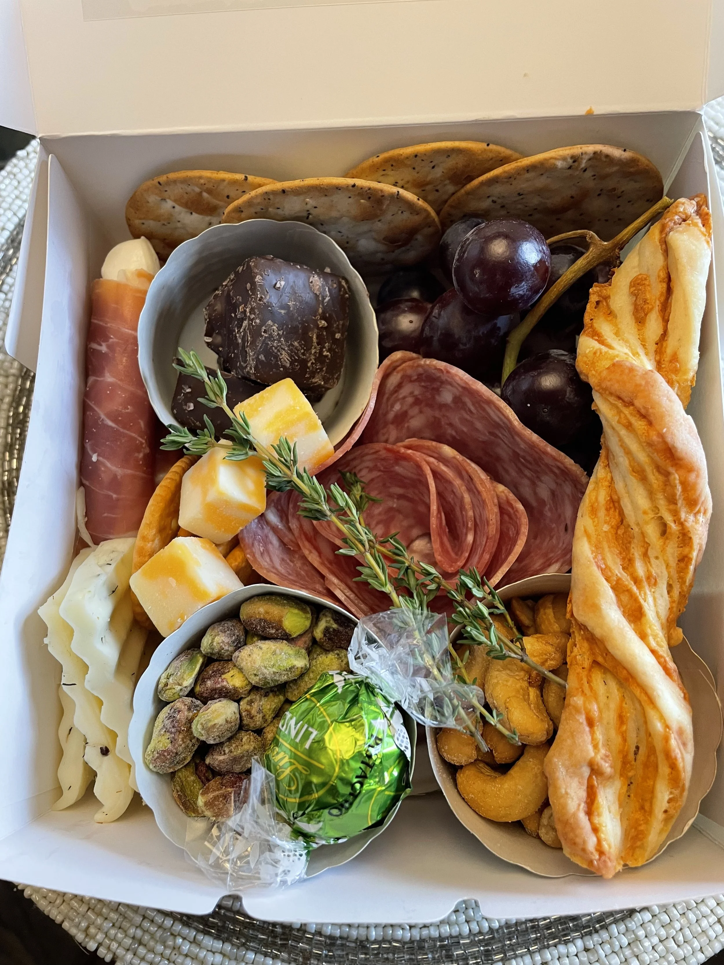 A white box with assorted snacks including cheese, cured meats, crackers, grapes, pistachios, and a small sprig of herbs.