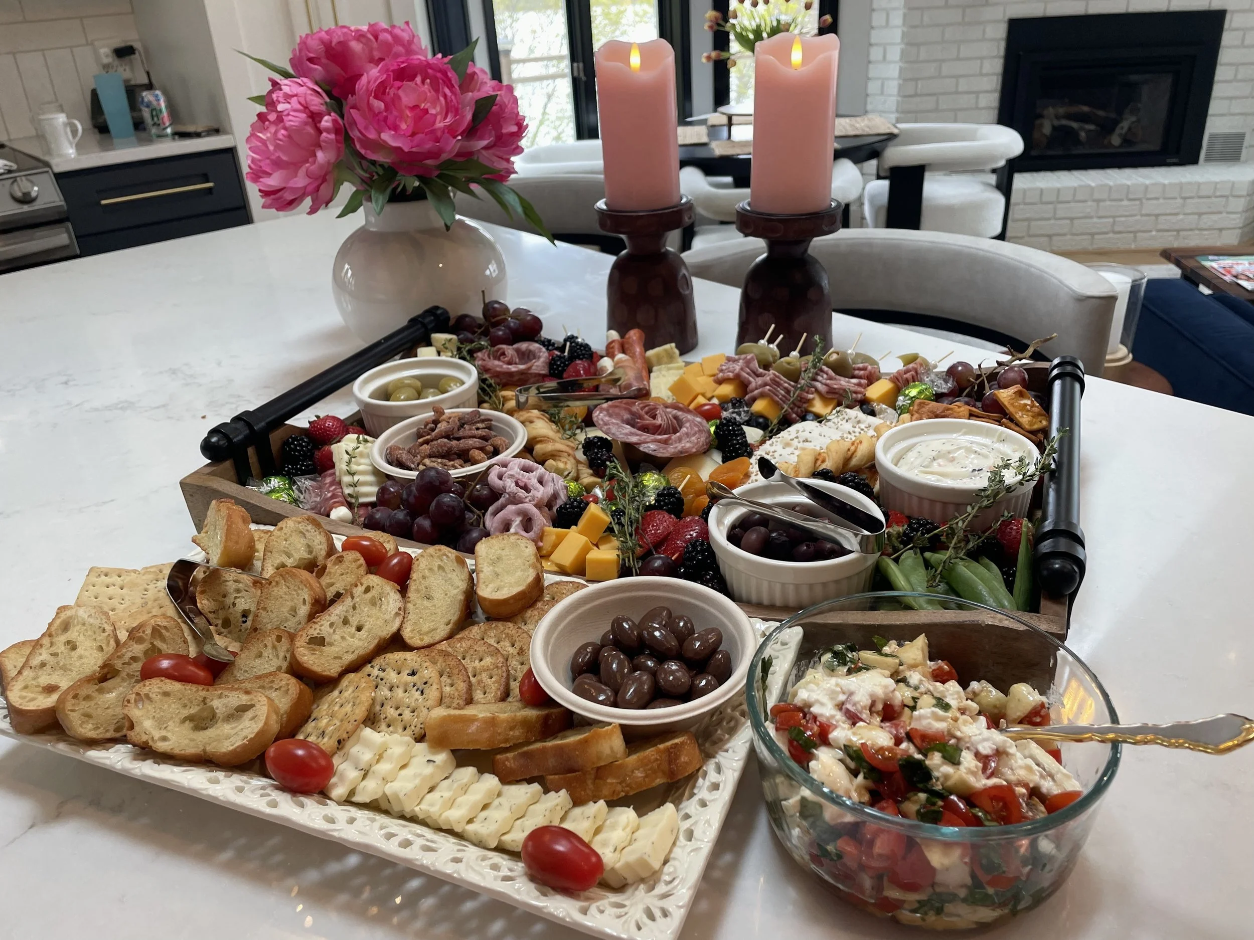 A tray of assorted appetizers, cheeses, fruits, and crackers, with a vase of pink peonies and lit pink candles on a white table in a cozy living room.