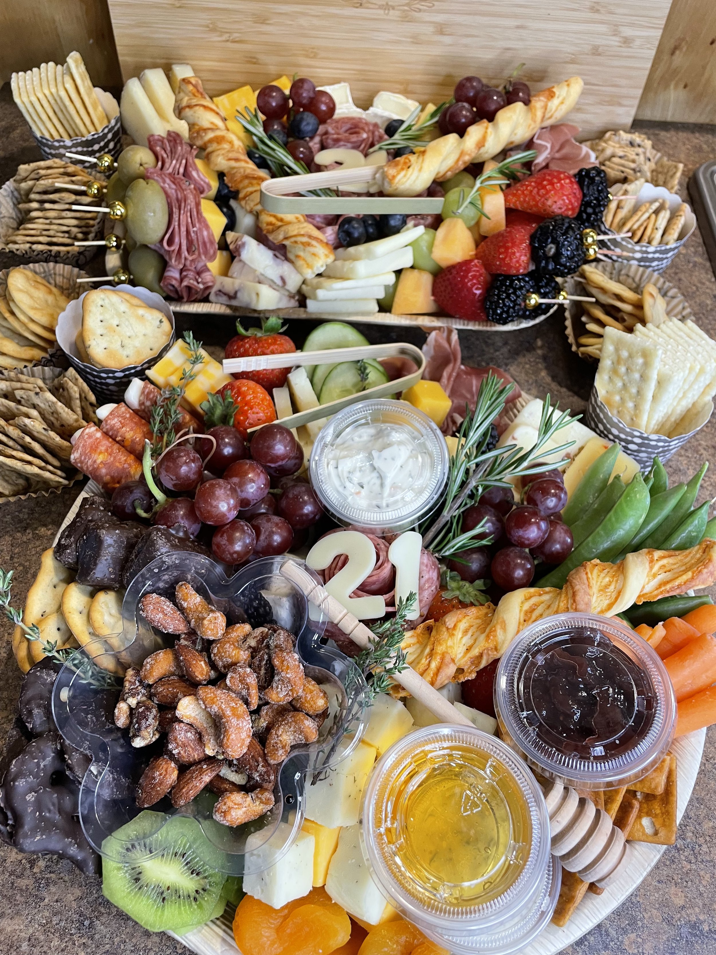 Assorted cheese, fruits, crackers, grapes, strawberries, blackberries, blueberries, orange slices, cucumber slices, and small containers of honey and jam on serving trays for a cheese platter.