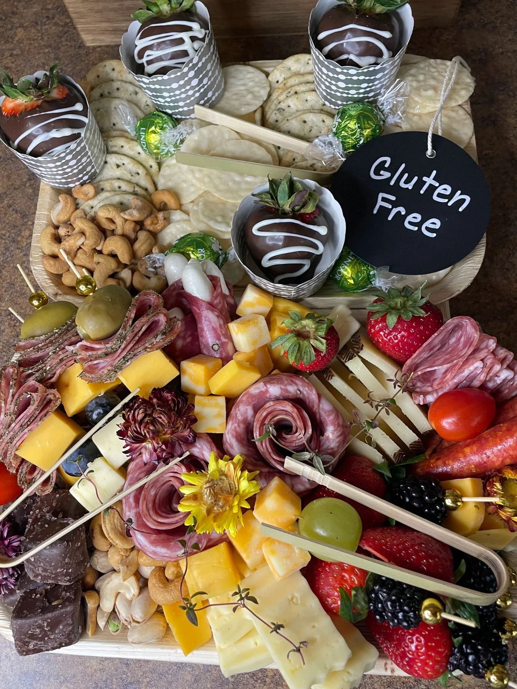 A cheese and fruit platter with strawberries, blackberries, grapes, cheese, cured meats, cashews, cookies, chocolate-covered strawberries, and decorative flowers, all arranged on a wooden tray with a black circular sign labeled "Gluten Free."