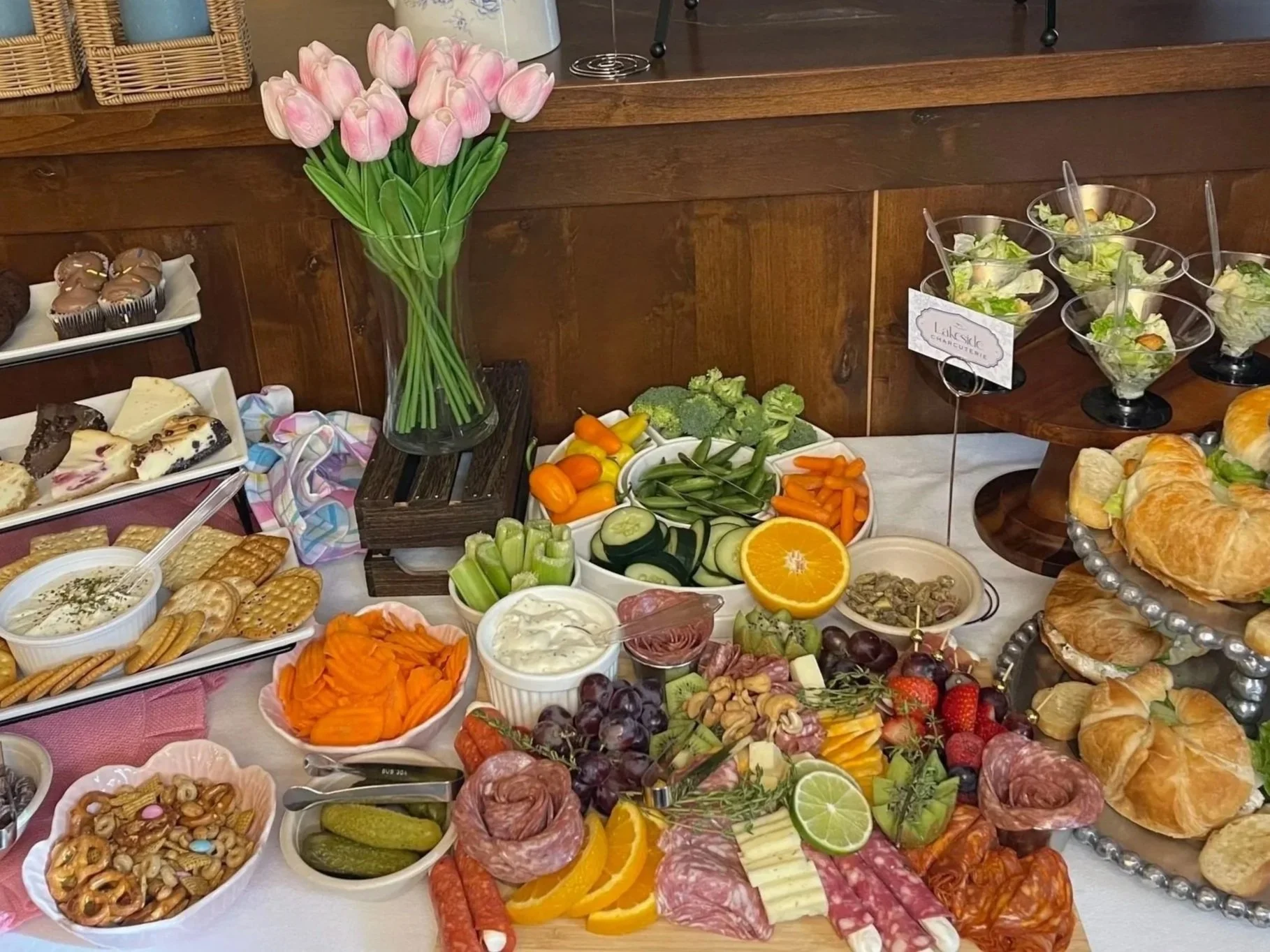 A table filled with various food items including fresh vegetables, fruits, cheese, meats, bread, and desserts, with a vase of pink tulips in the center.