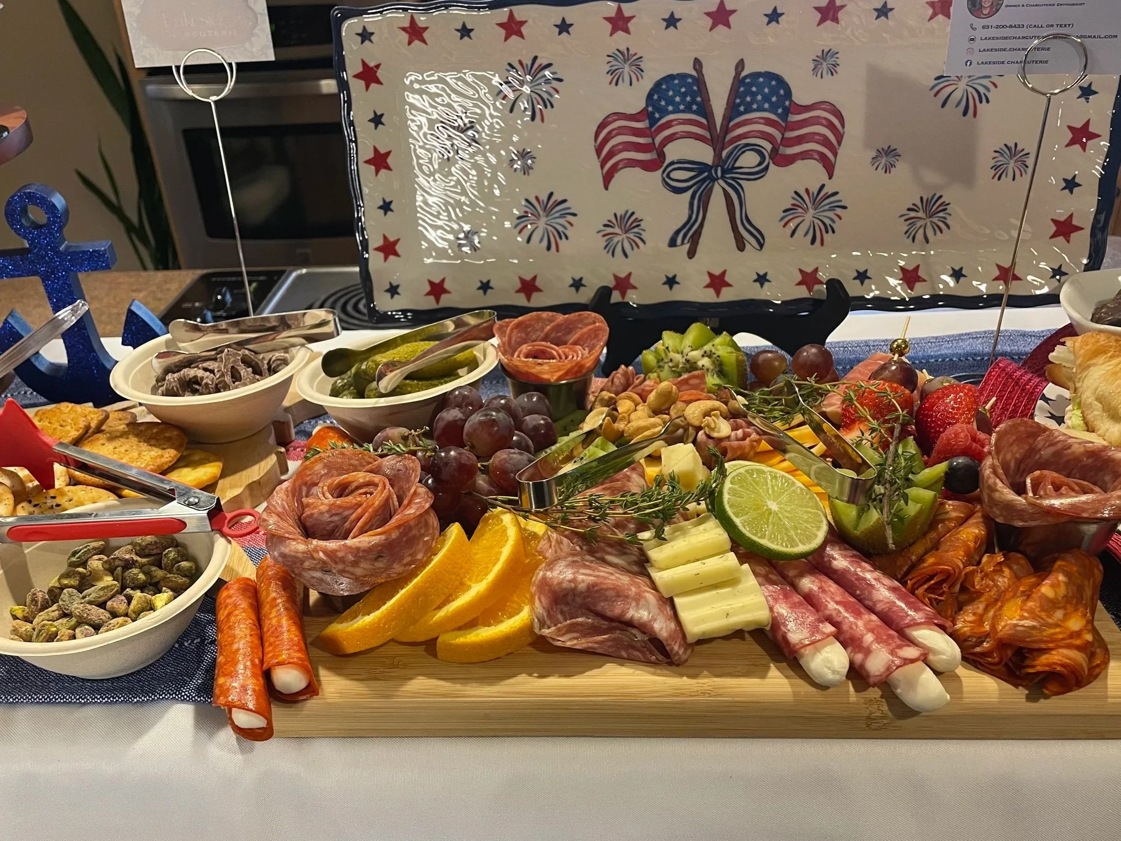 A patriotic holiday spread featuring an array of foods including sliced oranges, strawberries, grapes, green apple slices, cheese, cured meats, and crackers, with a large decorative plate in the background displaying red, white, and blue patriotic flags, stars, and fireworks.