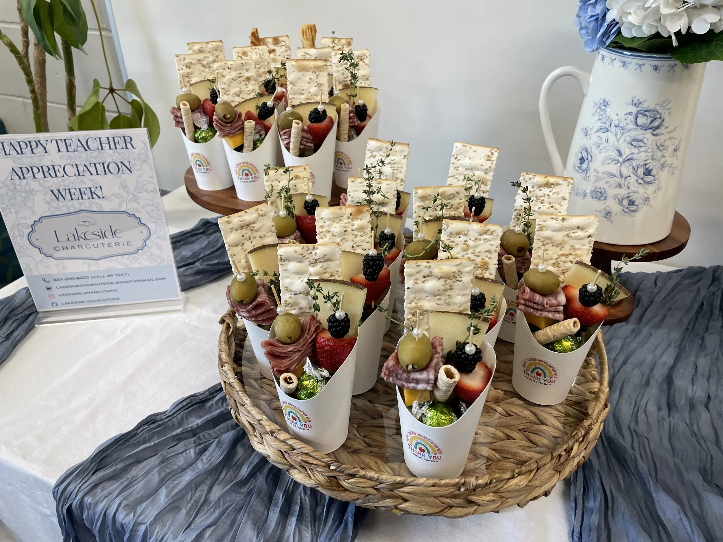 Assorted appetizers and snacks arranged in individual paper cones on a round woven basket, with a floral vase in the background, on a table.