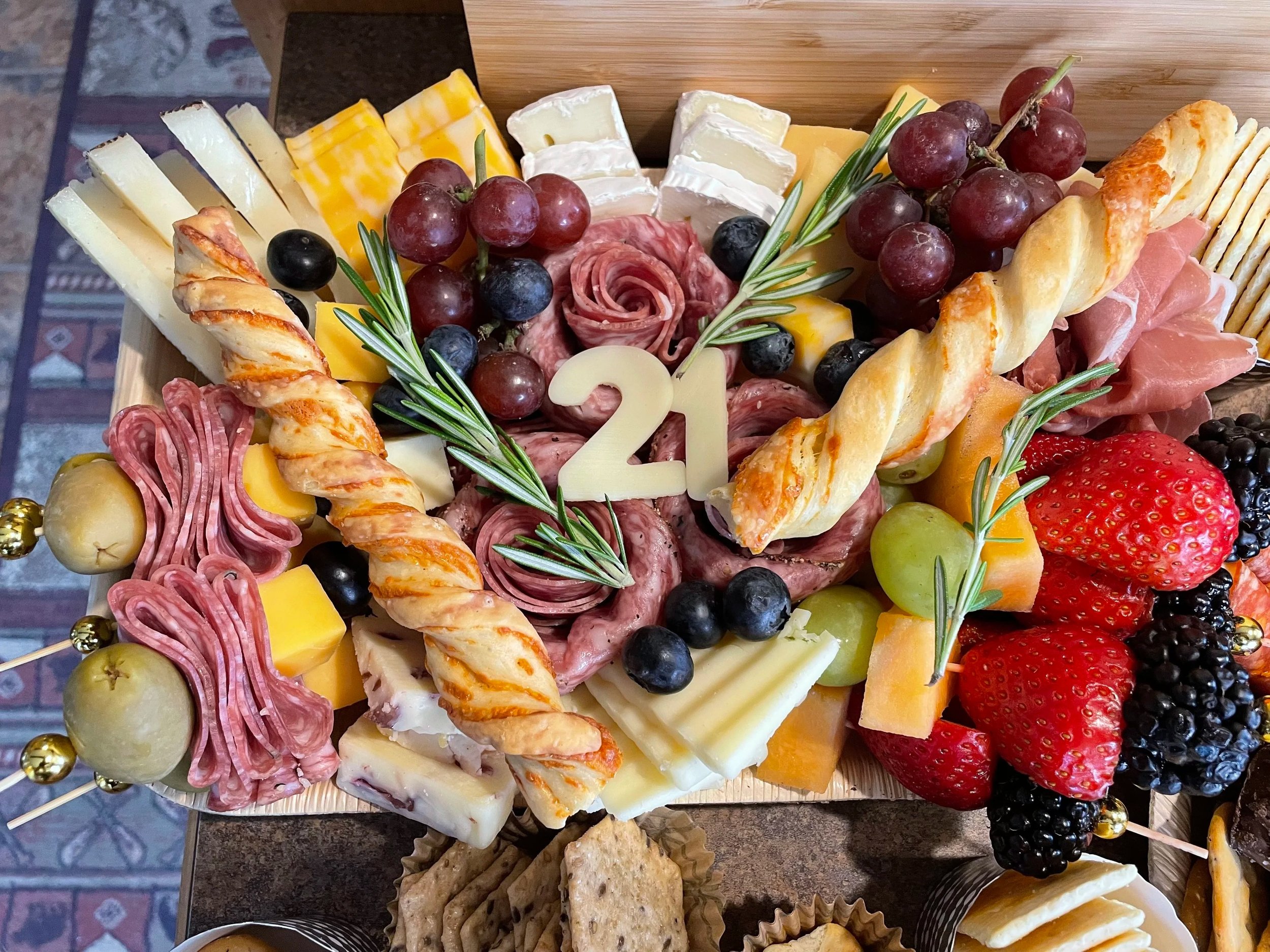 Cheese platter with slices of various cheeses, grapes, strawberries, blueberries, blackberries, sliced meats, crackers, and garnished with rosemary sprigs.