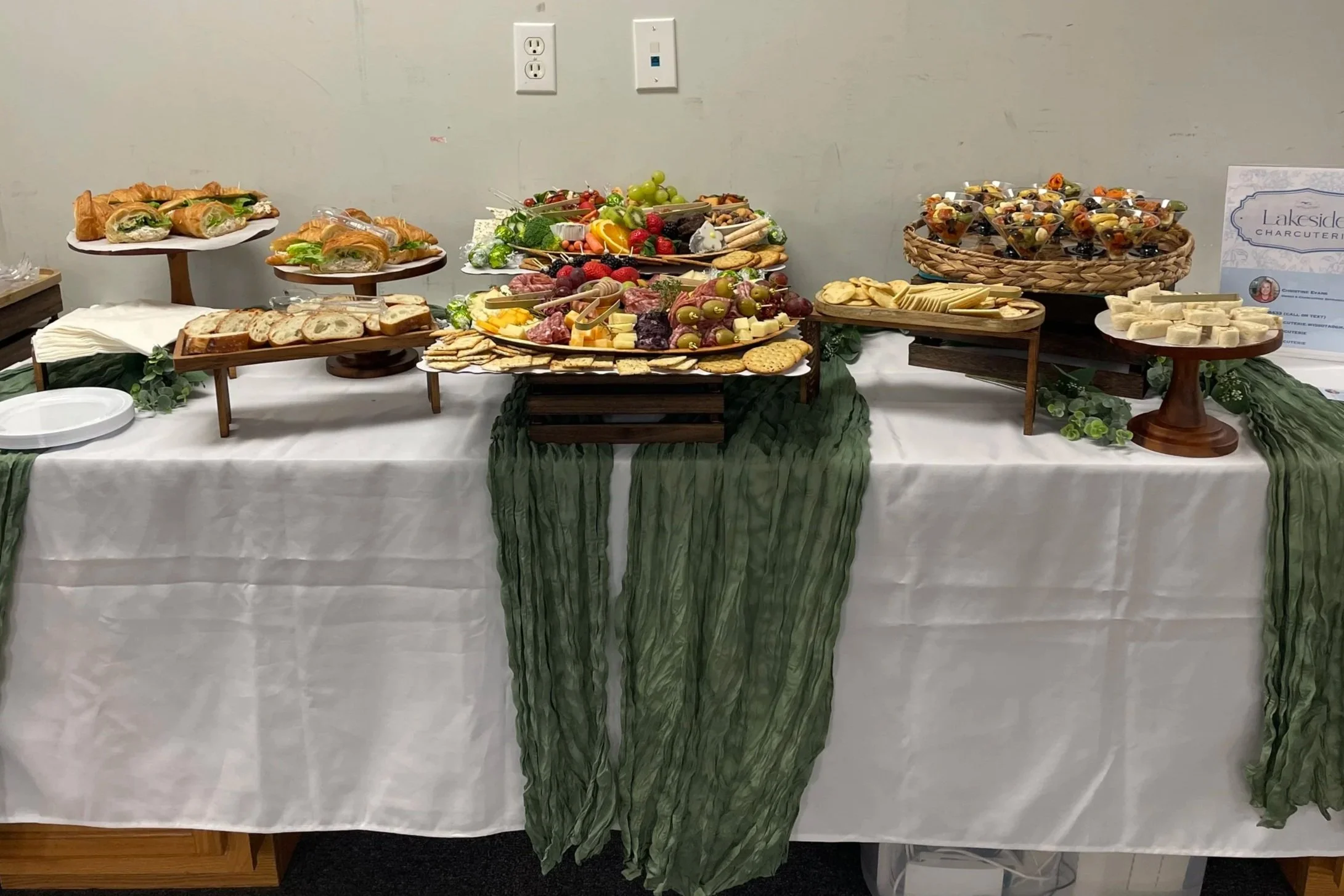 A buffet table with assorted food including croissant sandwiches, fresh fruit, cheeses, crackers, vegetable sticks, fruit cups, and a sign for Lakeside Charcuterie.