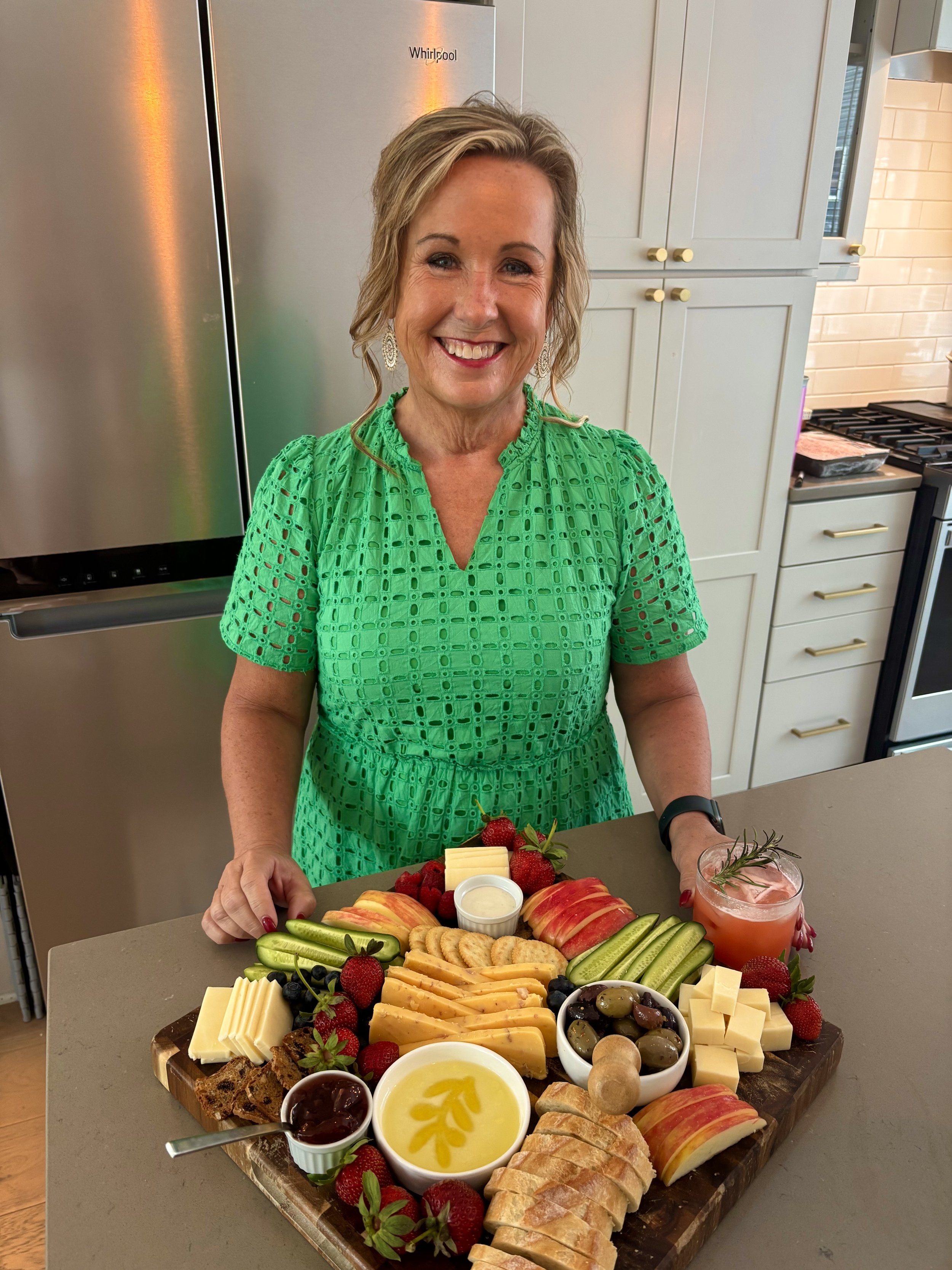 A woman in a green dress stands behind a large tray of assorted cheese, fruit, crackers, and toppings on a kitchen countertop.