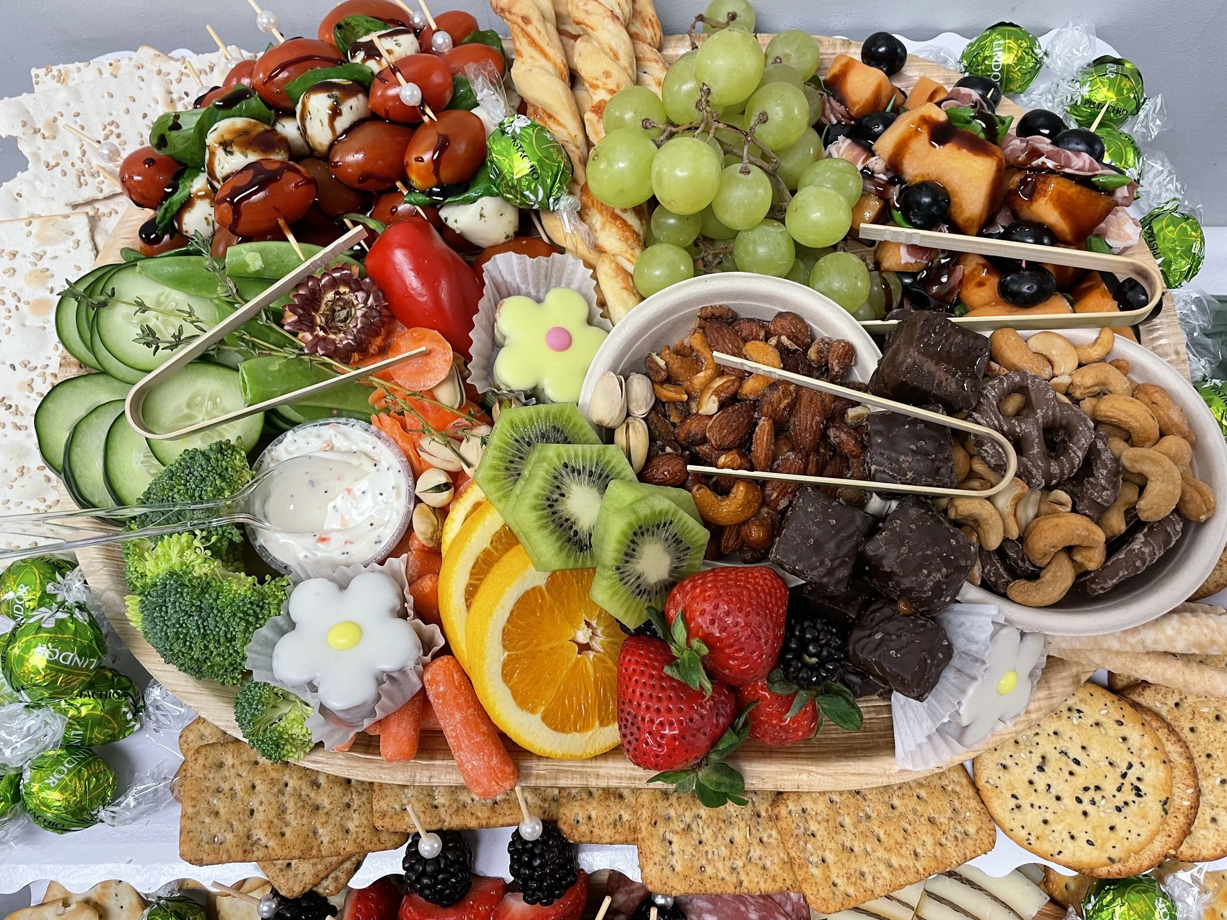 Assorted appetizer platter including grapes, strawberries, kiwi, vegetables, cheeses, crackers, chocolates, nuts, and candies.