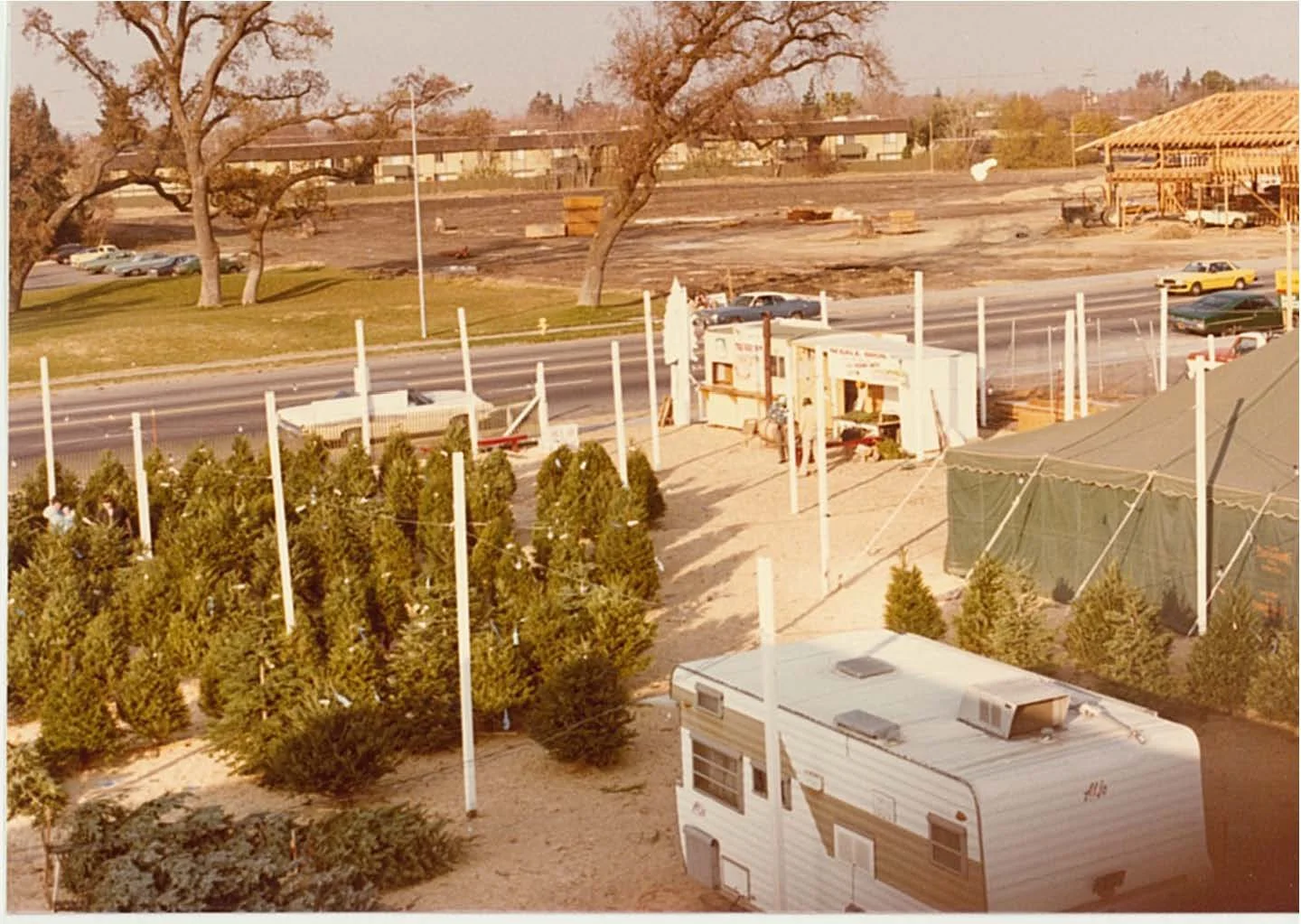 An outside scene showing a construction site, a street with parked and moving cars, some trees, and a small caravan in the foreground, with a makeshift booth or trailer near the trees.