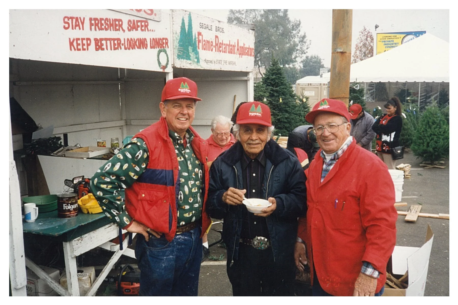 Three elderly men in front of a booth with Christmas trees, wearing red caps and red jackets, one holding a bowl, at an outdoor holiday event.