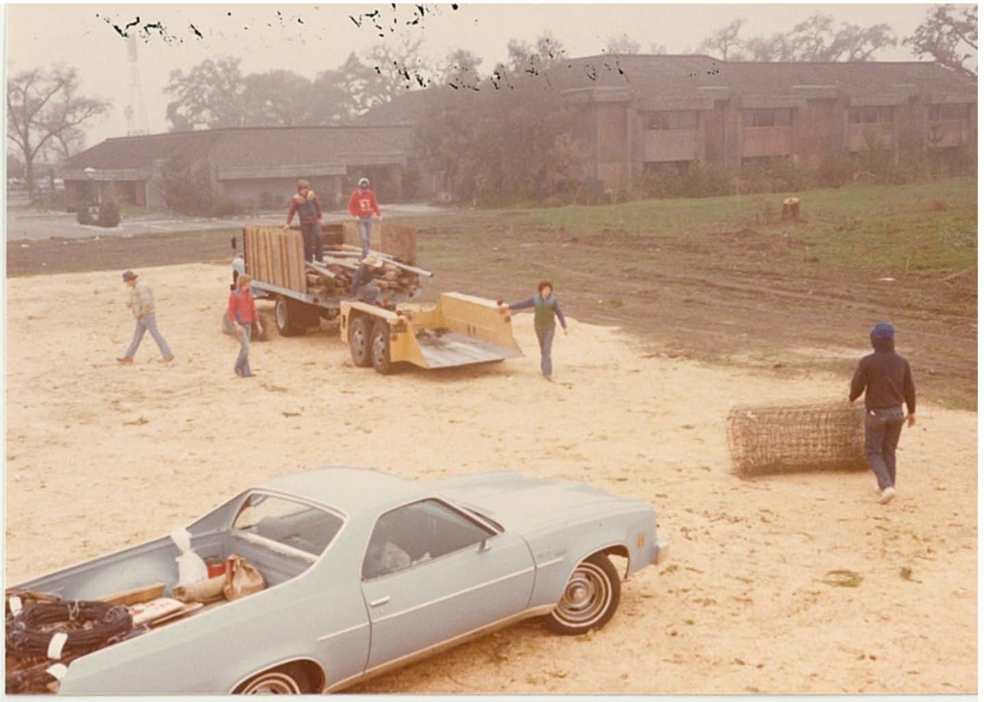 People working on a construction site, loading and moving items with a yellow bulldozer and a trailer, with a classic light blue car parked nearby and a building in the background.