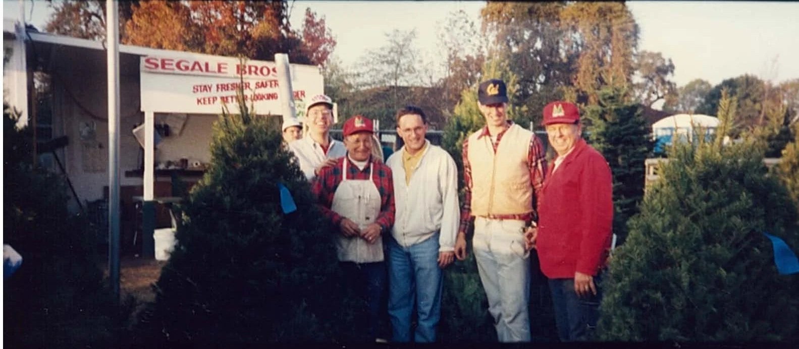Five people standing outdoors in front of greenery, some wearing Cubs hats, with a sign that reads 'SEGALE BROS STAY FRESH. SALT KEEP REFRIGERATED.'